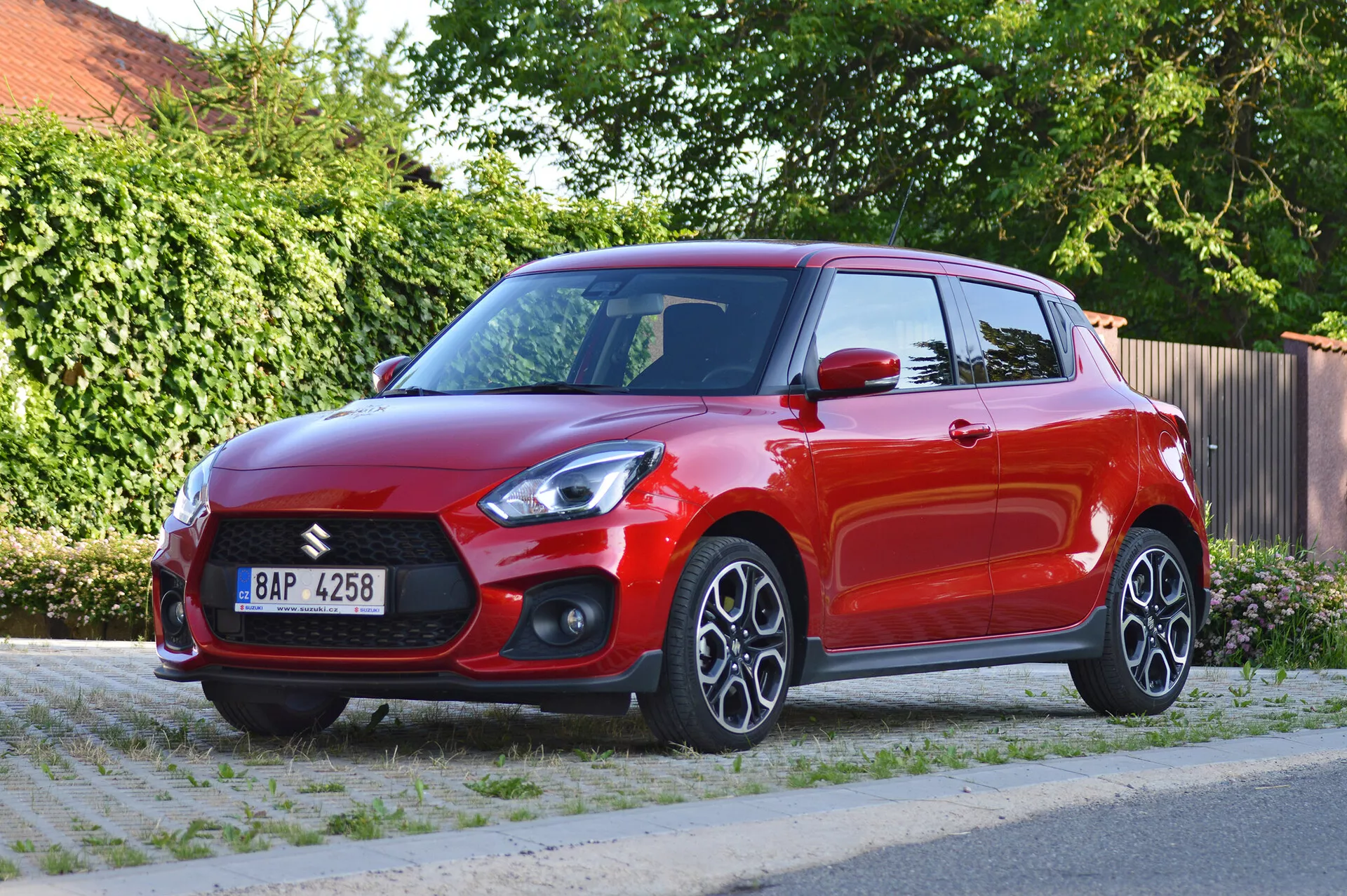 The image showcases a vibrant red 2017 Suzuki Swift Sport hatchback, photographed from a three-quarter frontal view. It highlights the prominent front grille with the Suzuki badge, sharp angular LED headlights, fog light housings, and air intakes in the lower front bumper. The hood features contour lines. The side profile shows multi-spoke alloy wheels, integrated turn signals on body-colored side mirrors, and body-colored door handles. The high shoulder line and slightly sloping roofline add to the sporty appearance. The glossy paint and slightly tinted windows indicate a well-maintained vehicle. The photo is taken during daylight with a clear view of a residential street and green foliage background, providing balanced exposure and emphasizing the car's design elements.