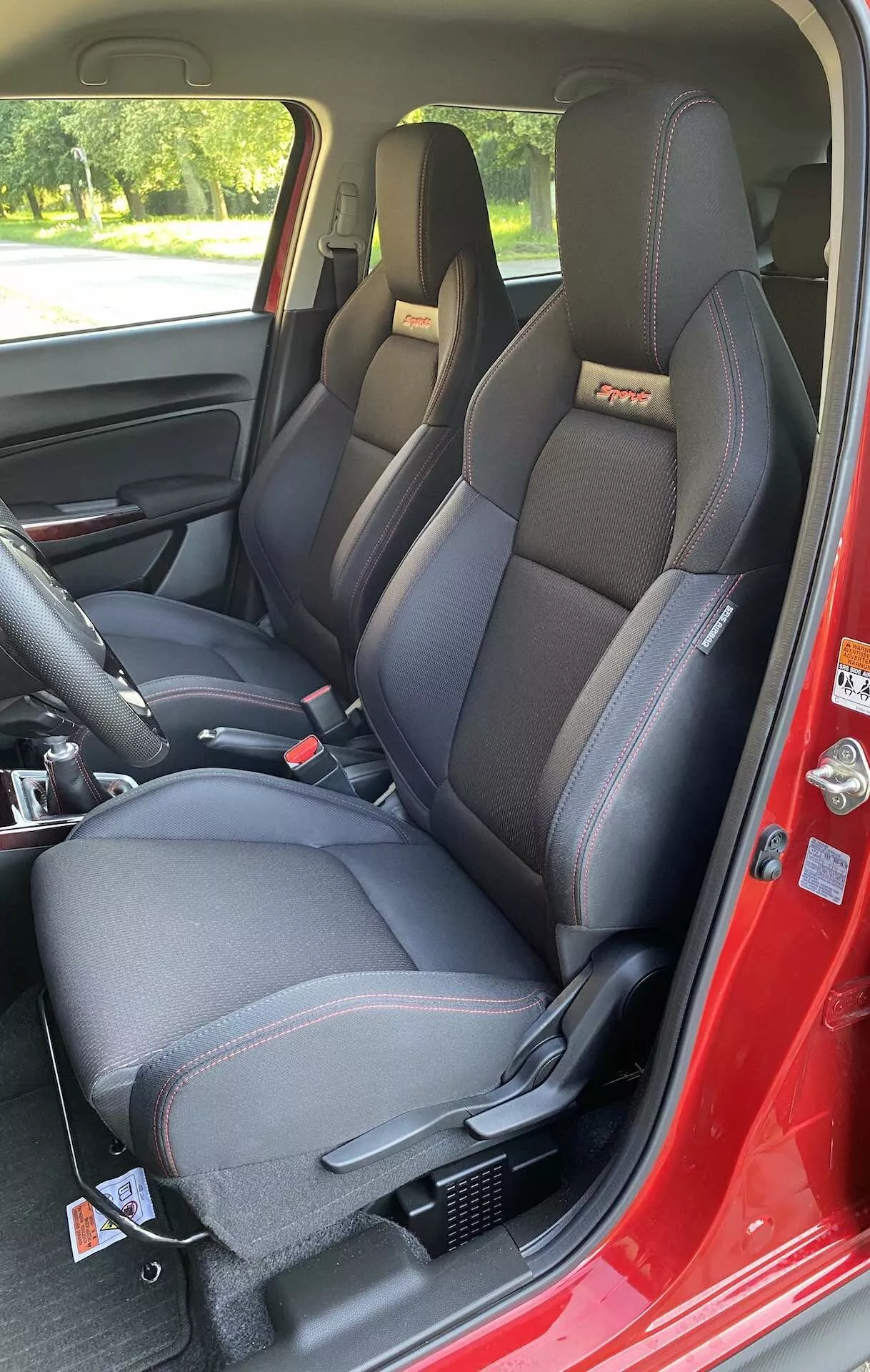 The interior of a 2017 Suzuki Swift Sport is shown from a passenger-side perspective through the open front passenger door. The focus is on the modern, sporty contoured front seats featuring integrated headrests, prominent side bolsters with a possible leatherette and fabric combination, and contrast stitching. Embossed or embroidered logo details are visible at the top of the backrests. Both seats are equipped with a three-point seat belt system, and the driver's side has an adjustable attachment for seat belt height on the B-pillar. The center console between the seats has storage compartments and a parking brake lever. The dark-colored interior door panel includes a door handle, mirror adjustment control, and power window controls. The floor is covered with dark carpet, and the base of the seats shows mounting rails. The daylight conditions and natural outdoor setting are visible through the windows, providing reflections and shadows that enhance the cabin's details.
