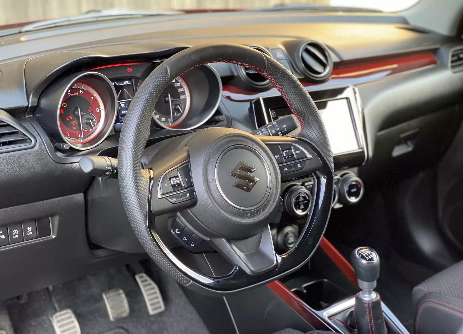 Interior view of a 2017 Suzuki Swift Sport from a slight right-side angle, featuring the multifunction leather-wrapped steering wheel, analog instrument panel with tachometer and speedometer, center console with multimedia touchscreen and manual controls, and gear shift knob for manual transmission.