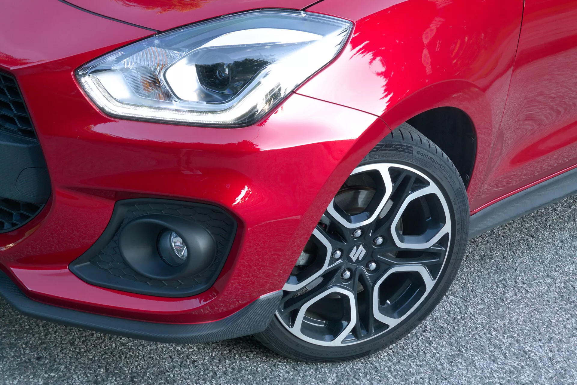 Close-up image showing the front left corner of the 2017 Suzuki Swift Sport from a downward and slightly frontward angle. The sleek, curved LED headlight emits a bright white light, suggesting a daytime running light feature. A small portion of the mesh-patterned front grille is visible below the headlight, adjacent to a circular element that could be a fog lamp or parking sensor set within the dynamic, sculpted front bumper. The matte black section of the bumper contrasts with the bright red paintwork of the car. The multi-spoke alloy wheel design with dark and silver elements stands out, with tire tread visible, indicating it is in good condition. The car's low ride height is noticeable, reflecting a standard ride height suitable for a sporty model.