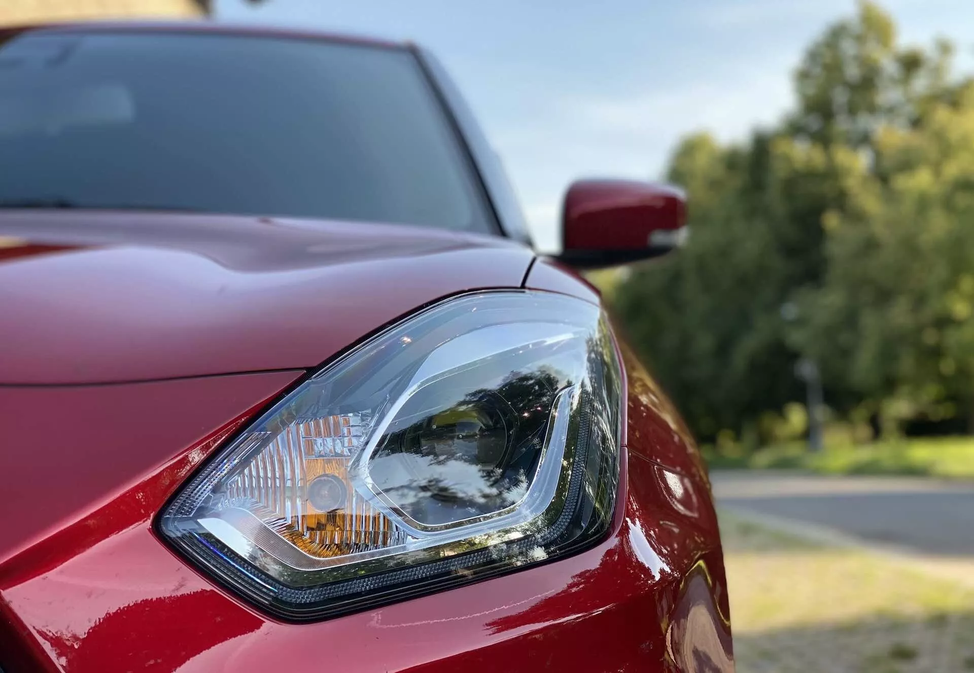 Close-up of the driver's side headlight on a Suzuki Swift Sport 2017. The image is taken from the front quarter, highlighting the design details of the clear lens headlight with its complex internal reflector and amber turn signal indicator. Visible are glossy, deep red body panels including the hood and fender, set against a soft-focus outdoor background with trees and a bright sky.