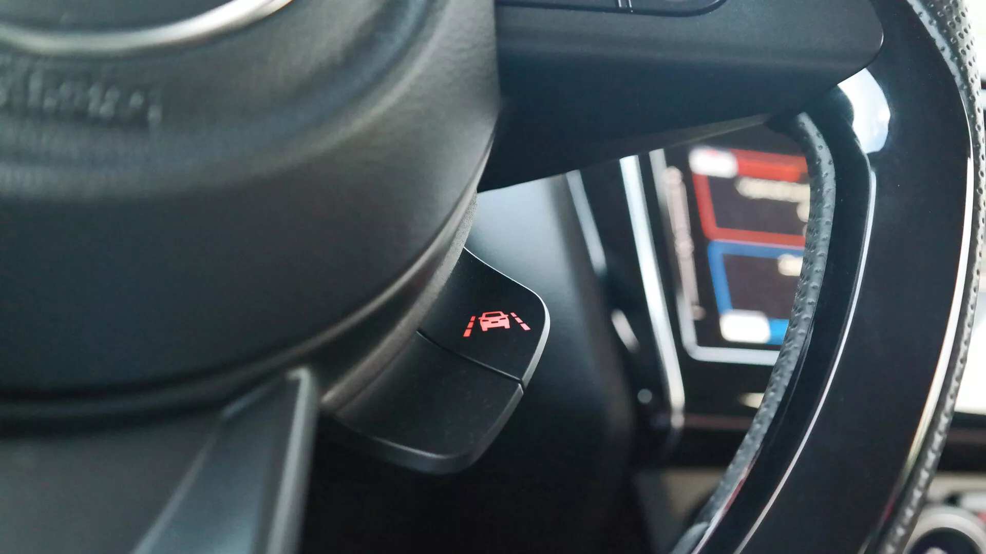 Interior view of a 2017 Suzuki Swift (Swift-sport) from the driver’s seat perspective, focusing on the steering wheel and control stalk. The dark-colored, possibly leather, steering wheel features a metallic or glossy trim. A control stalk for cruise control with a gauge and arrow symbol protrudes from the steering column. In the blurred background, the digital instrument cluster displays various numbers and lines, indicating advanced technological features. The interior materials show a mix of synthetic or leather textures with intricate stitching, and the shallow depth of field highlights the control stalk with the dashboard in soft focus.