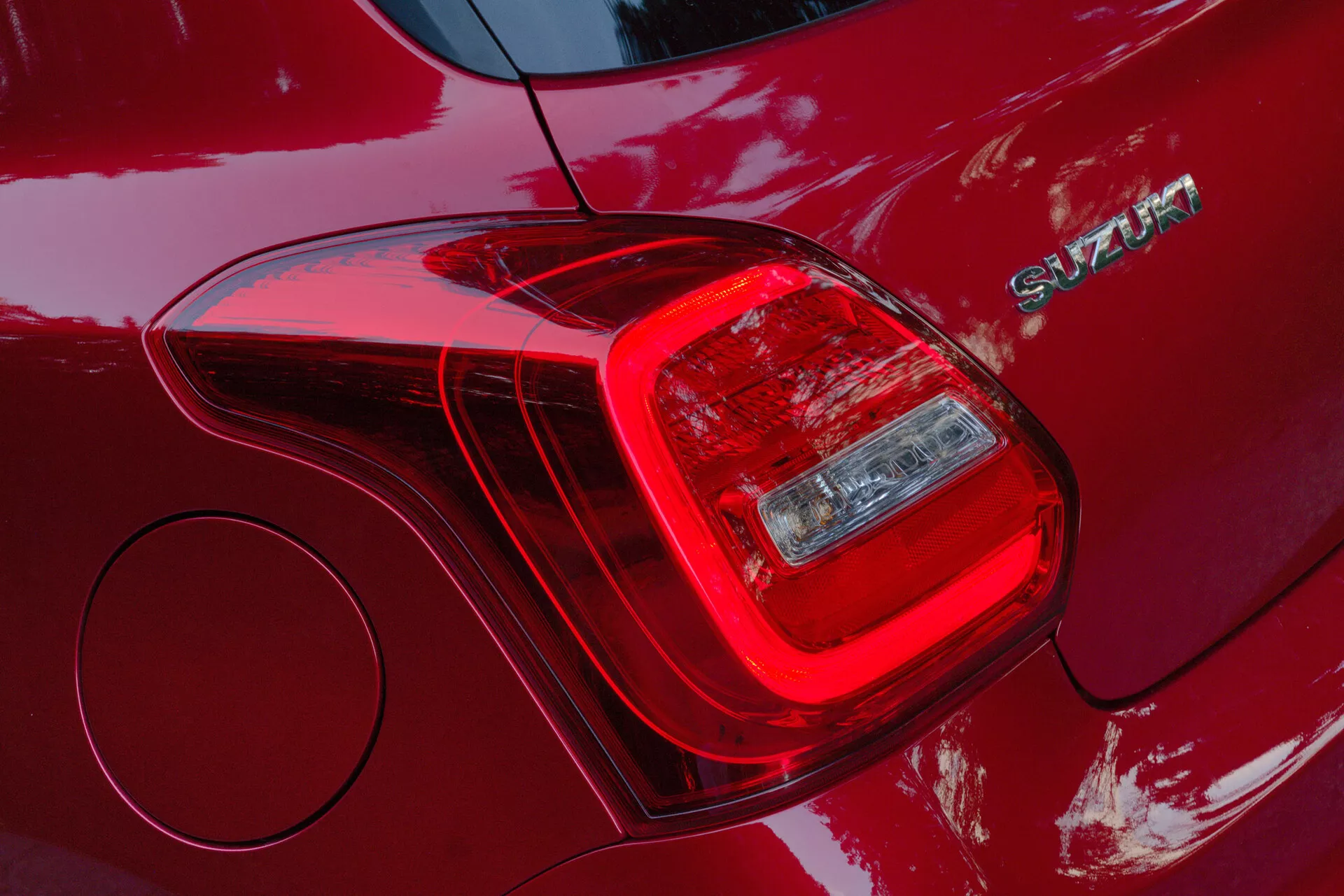 Close-up of the rear left taillight assembly of a 2017 Suzuki Swift Sport, highlighting the segmented design of the light units including brake light and turn signal or reverse light. The deep red, glossy body paint reflects light and surroundings. Branding with a chrome 'SWIFT' emblem is visible beside the taillight. The shot also reveals a circular fuel cap cover and showcases a sleek and modern design language with aerodynamic lines and durable construction materials.