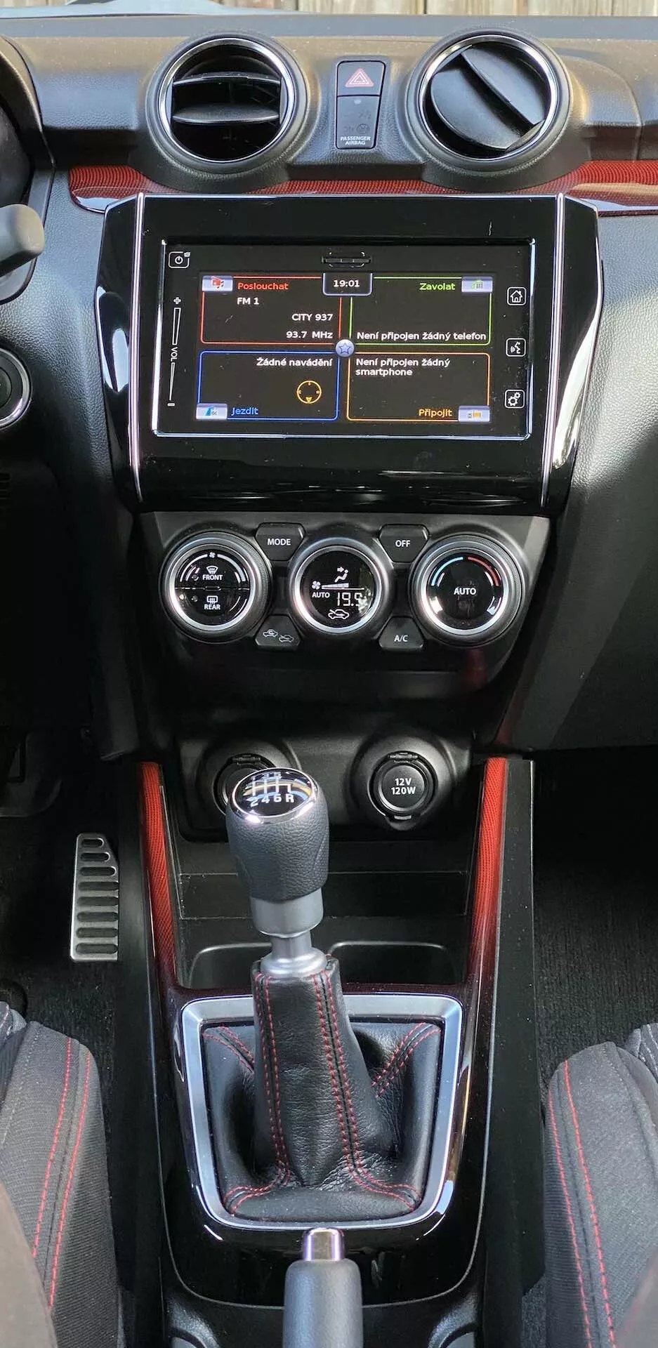 The interior center console of a 2017 Suzuki Swift Sport, featuring an infotainment system with physical buttons, circular air vents, a manual climate control panel with three dials for temperature, fan speed, and air distribution, and a sporty manual gear shift knob. The console includes a leather boot, decorative trim with a possible carbon fiber-like pattern, and a sporty red accent line. Additional controls below the gear shift hint at advanced driving aids.