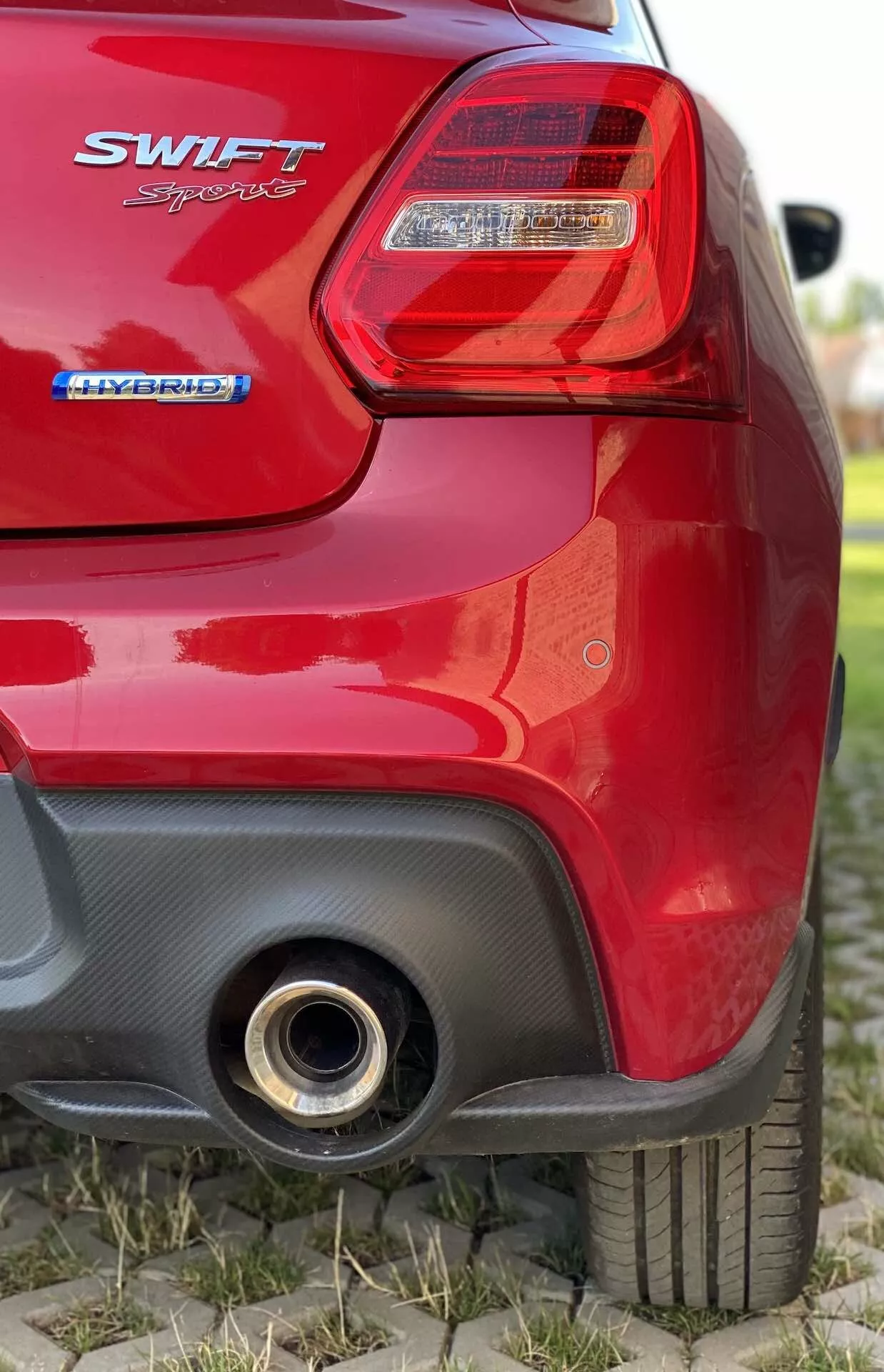 Close-up of the rear section of a red Suzuki Swift 2017 hatchback from a low angle. Image focuses on the right tail light with clear lens and red illumination sections, 'SWIFT' badge below the light, 'HYBRID' badge underneath it indicating hybrid powertrain, partially out-of-focus rear hatch, back right tire with visible tread pattern, black contrasting section on the rear bumper with possible aerodynamic intake, and single round metallic exhaust pipe on the bottom right side reflecting light.