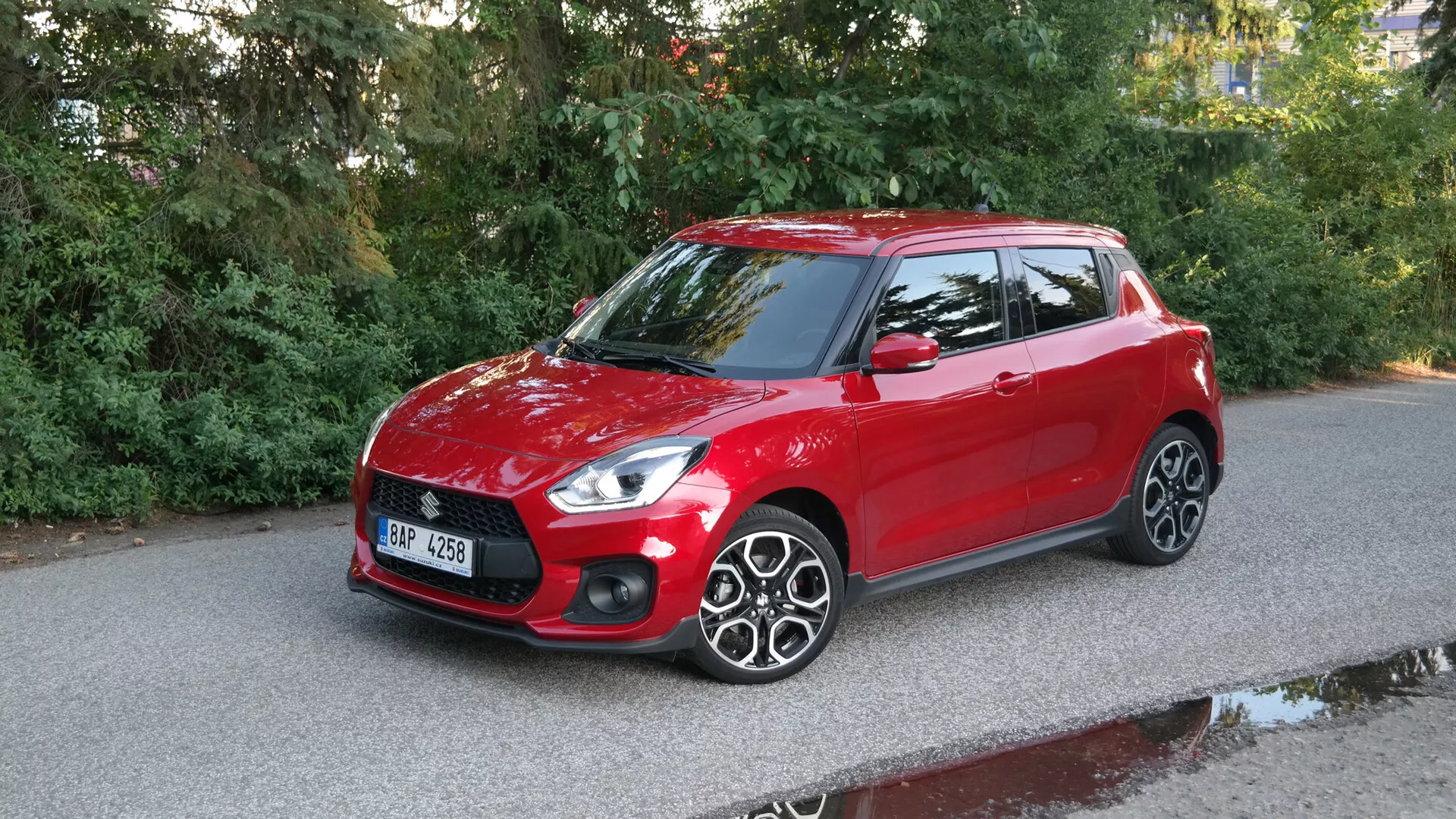 Image of a red 2017 Suzuki Swift Sport hatchback photographed from a three-quarter front angle, showcasing the sleek headlights, prominent front grille with horizontal lines, smooth hood, clear windshield, driver's side including body-colored side mirror, multi-spoke alloy wheels, and roof spoiler against a paved road with natural surroundings.