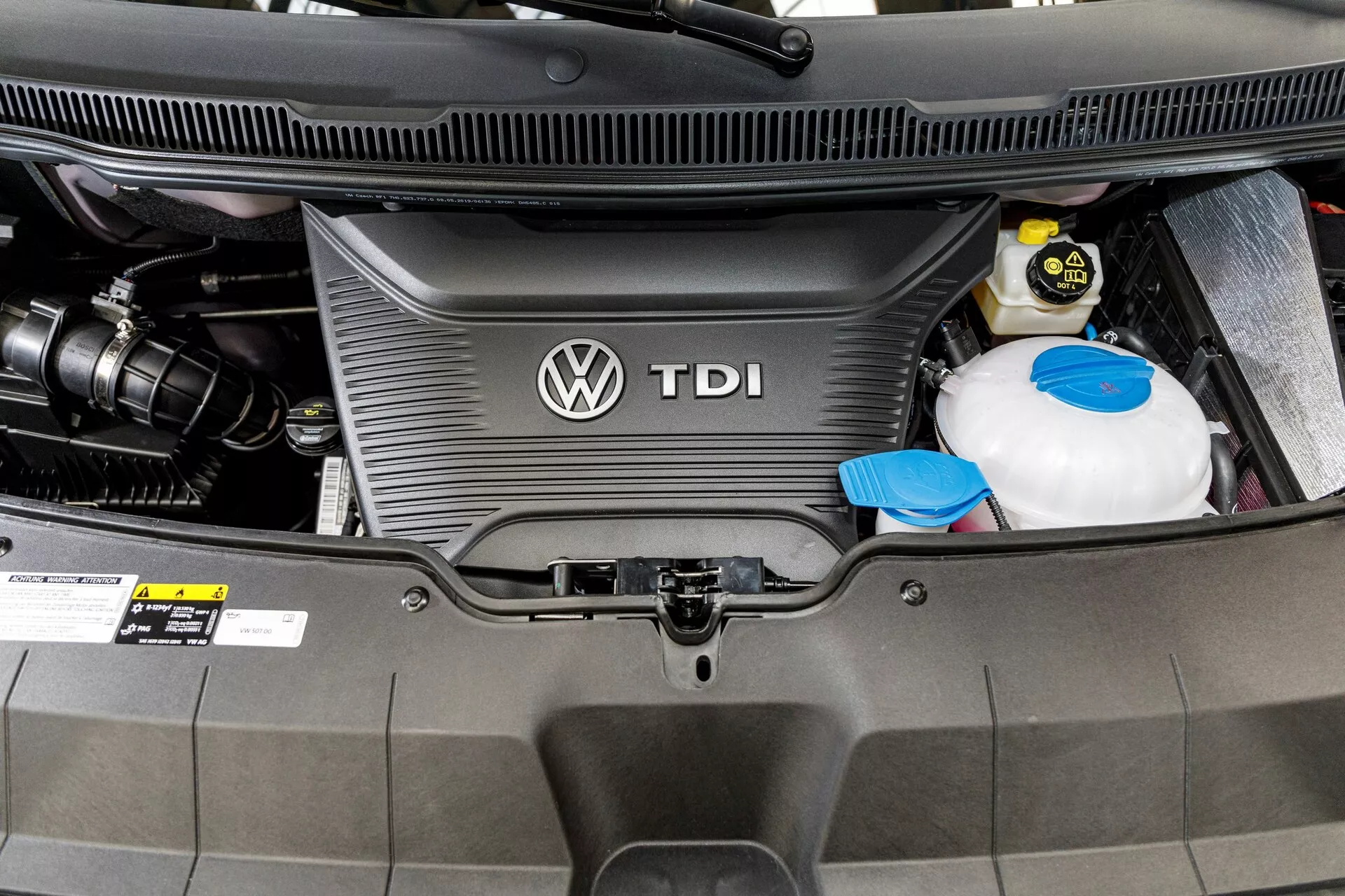 Close-up of VW Nutzfahrzeuge Transporter 2019 (T6.1 California) diesel engine bay showing TDI engine cover with Volkswagen logo, coolant reservoir, and car battery