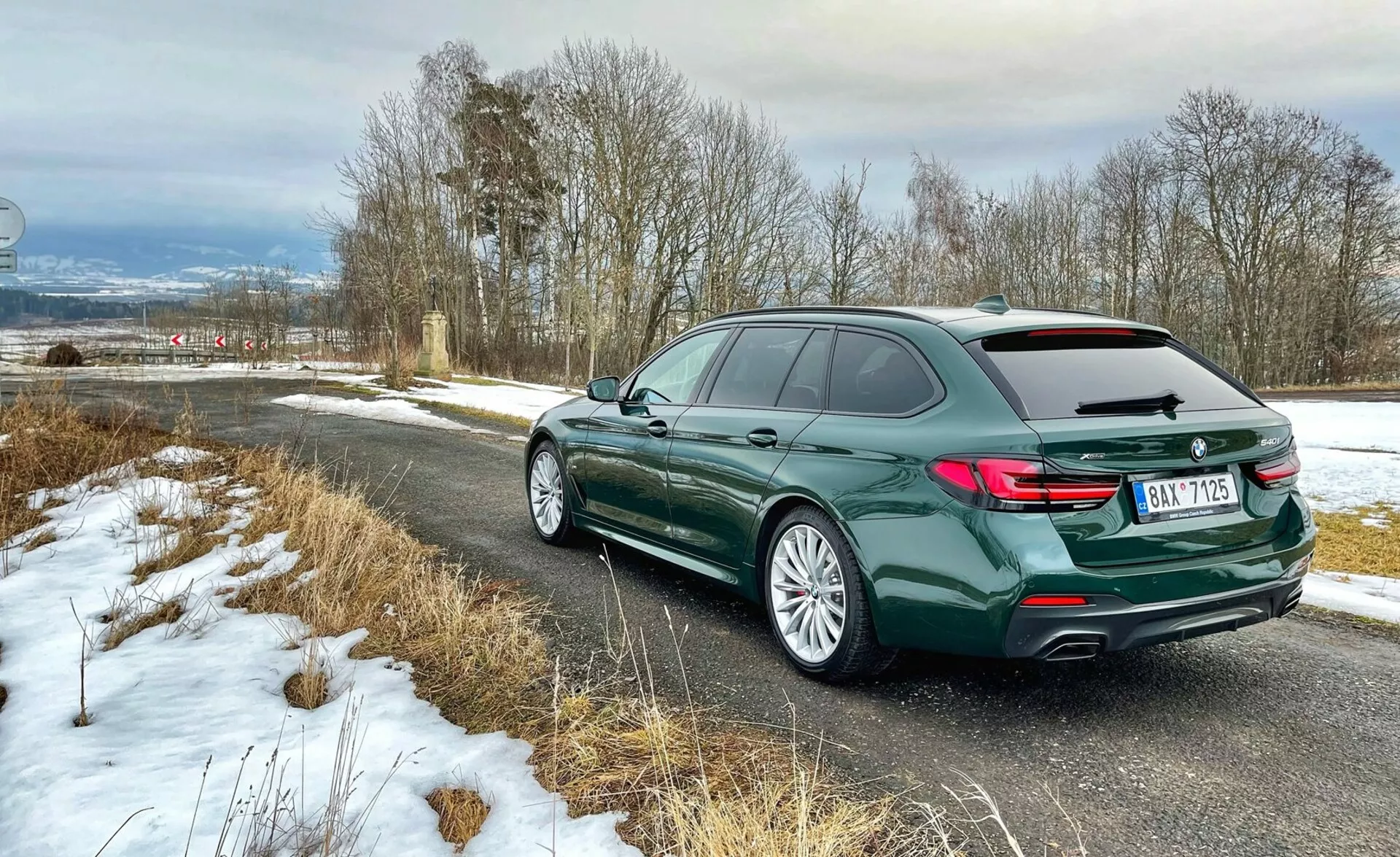 Rear three-quarter view of a BMW station wagon, showcasing the vehicle's rear end with L-shaped taillights, the left side profile including doors and roof curve with roof rails, alloy rims with road tires, in a deep green metallic paint, parked on a country road with light snow, highlighting luxury and modern design elements.