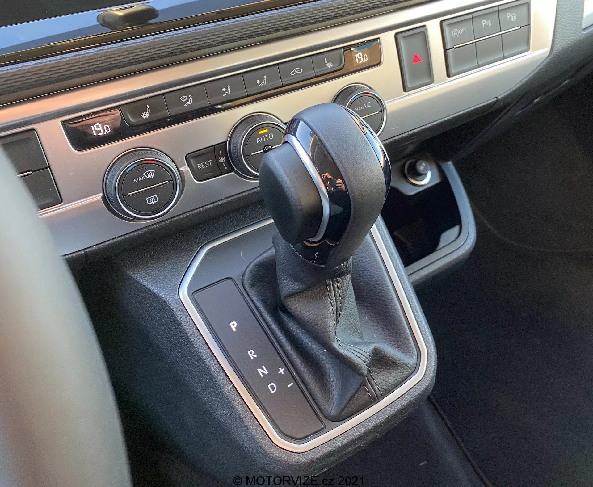 Close-up view of the center console of the 2019 VW Nutzfahrzeuge T6-1 Multivan focusing on the gear shift area. The image shows an automatic transmission selector with leather stitching, surrounded by metallic trim, climate control buttons with temperature display, and seat heating controls, showcasing the vehicle's modern interior technology and premium materials.