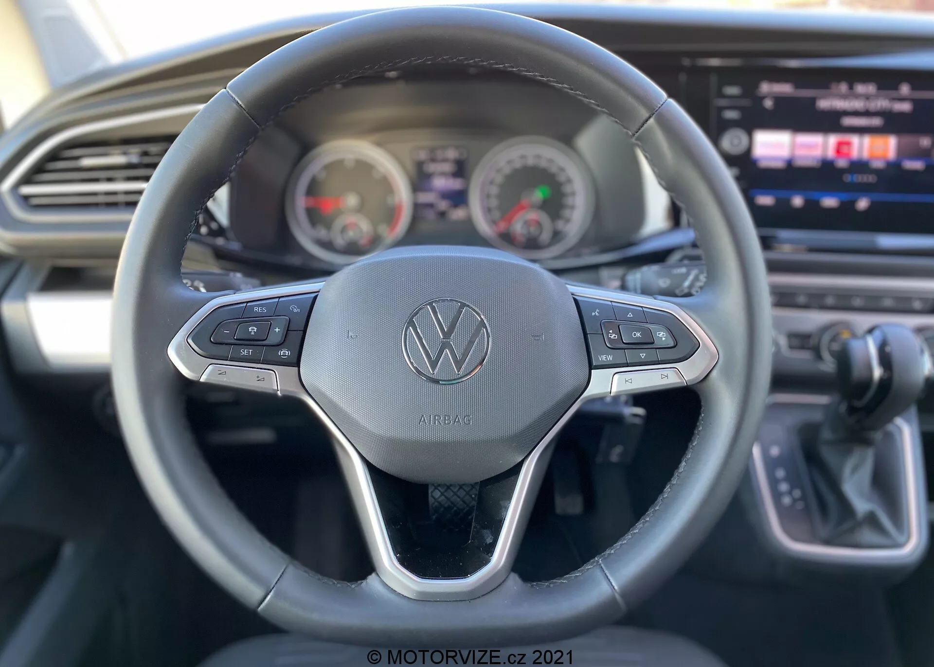 Interior view of a 2019 VW Nutzfahrzeuge Transporter (T6-1 Multivan) focusing on the driver's perspective. Detailed view of the steering wheel featuring the Volkswagen logo with control buttons for various functions like cruise control and multimedia navigation. Behind the wheel, the instrument cluster shows analog-style speedometer and tachometer with a digital display in between. Partially visible center console housing a large infotainment touch-screen display, alongside sleek dashboard design with horizontal air vent. Gear shift lever indicating automatic transmission to the far right. The image highlights usability features with shallow depth of field primarily focusing on the steering wheel.