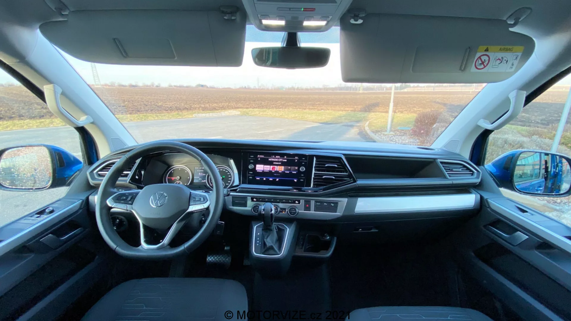 Interior of a 2019 VW Nutzfahrzeuge Transporter (T6.1 Multivan), captured from the rear seating area looking towards the front dashboard. The photo is taken during the day with natural light entering through the front windshield, providing a detailed view of the cabin space, front seats, steering wheel with mounted controls, central infotainment touchscreen display, HVAC buttons and knobs, automatic gear shifter, storage spaces, connectivity ports, door and mirror controls, safety features, and comfortable seat design.