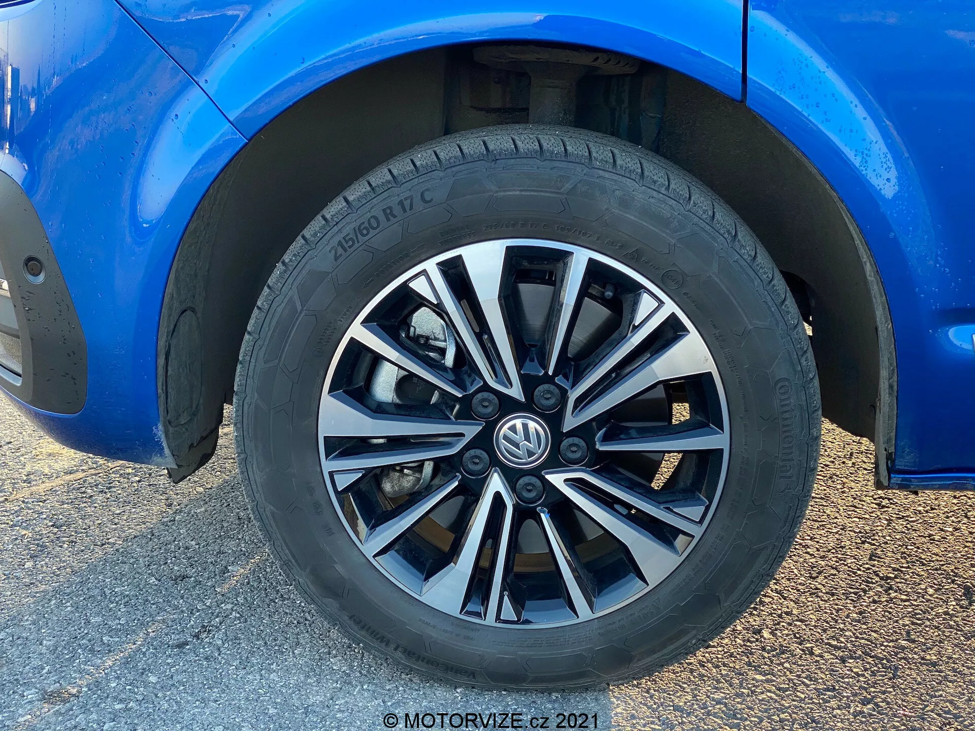 Close-up view of a Volkswagen T6-1 Multivan 2019 wheel. This full-frame shot features a multi-spoke design wheel with alternating silver and dark finishes. The tire, branded Hankook, reveals a street performance tread pattern and partial tire specifications. The surrounding blue car body, in pristine condition, shows a reflective sheen. A moderate ground clearance and partially visible standard brake calipers and rotors are indicated. Natural light enhances the wheel-focused composition, with the background blurred due to a low-angle close-up shot.
