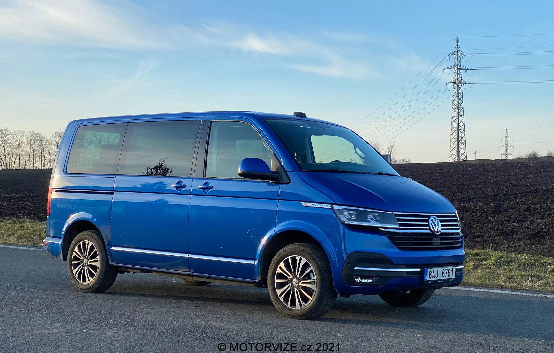 A blue Volkswagen Transporter T6 van, showing its side profile from a low angle, emphasizing its length and height. The image features details such as the sliding passenger door, front passenger door, rear passenger-side wheel, multi-spoke alloy rims, and partial views of the headlight, Volkswagen grille with emblem, and front bumper. The photo, taken in the late afternoon, showcases ground clearance and reflects sky and surroundings in the windows, with a clear sky and power lines in the background.