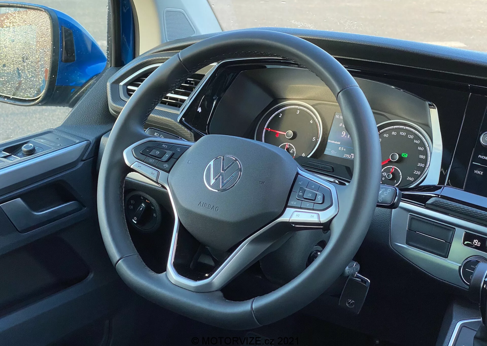 Interior view of the 2019 VW Nutzfahrzeuge Transporter (T6-1 Multivan) featuring the driver's seat perspective with a focus on the steering wheel and dashboard. The steering wheel showcases the Volkswagen emblem and is equipped with leather wrapping, multifunctional buttons, and paddle shifters. The partially visible instrument cluster includes analog dials and a digital screen. Central infotainment screen and climate controls are faintly visible, indicating modern multimedia and comfort features.