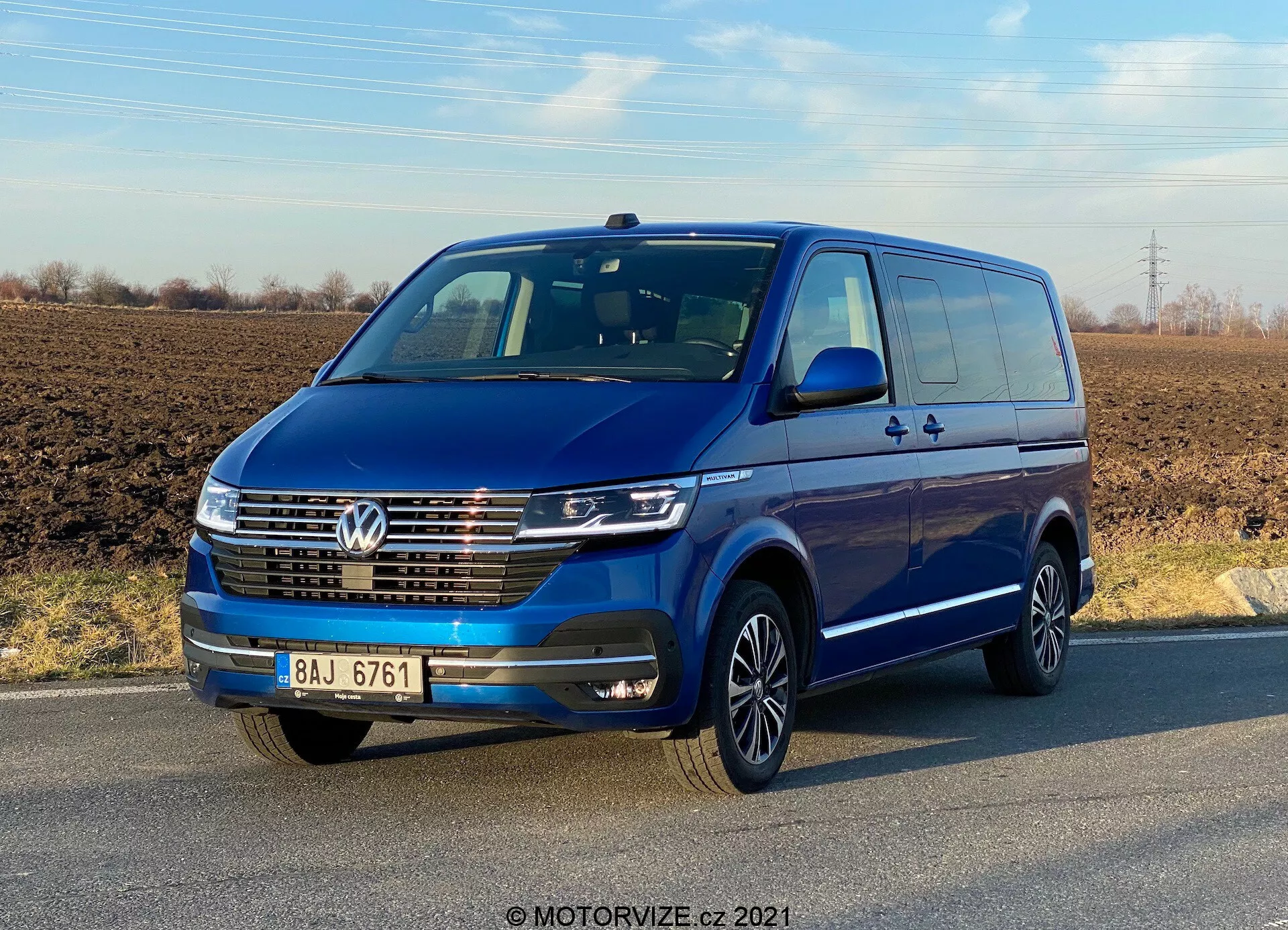 A three-quarter front view of a blue 2019 Volkswagen Transporter T6.1 Multivan under a clear sky. The vehicle features a Volkswagen badge on the grille, LED daytime running lights, chrome trim on the front grille, and multi-spoke alloy wheels. Body-colored side mirrors and bumpers suggest a high-spec model. Doors and door handles are visible including a rear sliding door, indicating its capacity for multiple passengers or cargo.