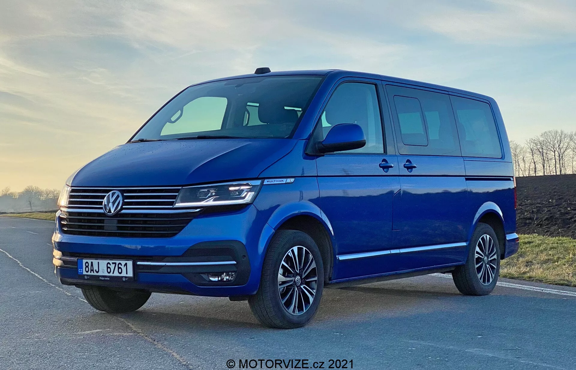 Image of a blue 2019 Volkswagen Transporter T6.1 Multivan, shot from a low angle to accentuate its stature and size. The front three-quarter view showcases the vehicle's frontal and lateral design, including the distinctive Volkswagen grille with horizontal lines, the central emblem, modern headlights likely featuring LED or Xenon technology, and a sleek front bumper with a wide air intake. The streamlined side mirrors with integrated turn signals, ample window space with a sliding side door, alloy rims with low-profile tires, and soft lighting highlight the van's practicality and aesthetic appeal. The watermark 'MOTORVIZE.cz 2021' suggests the image is from 2021.
