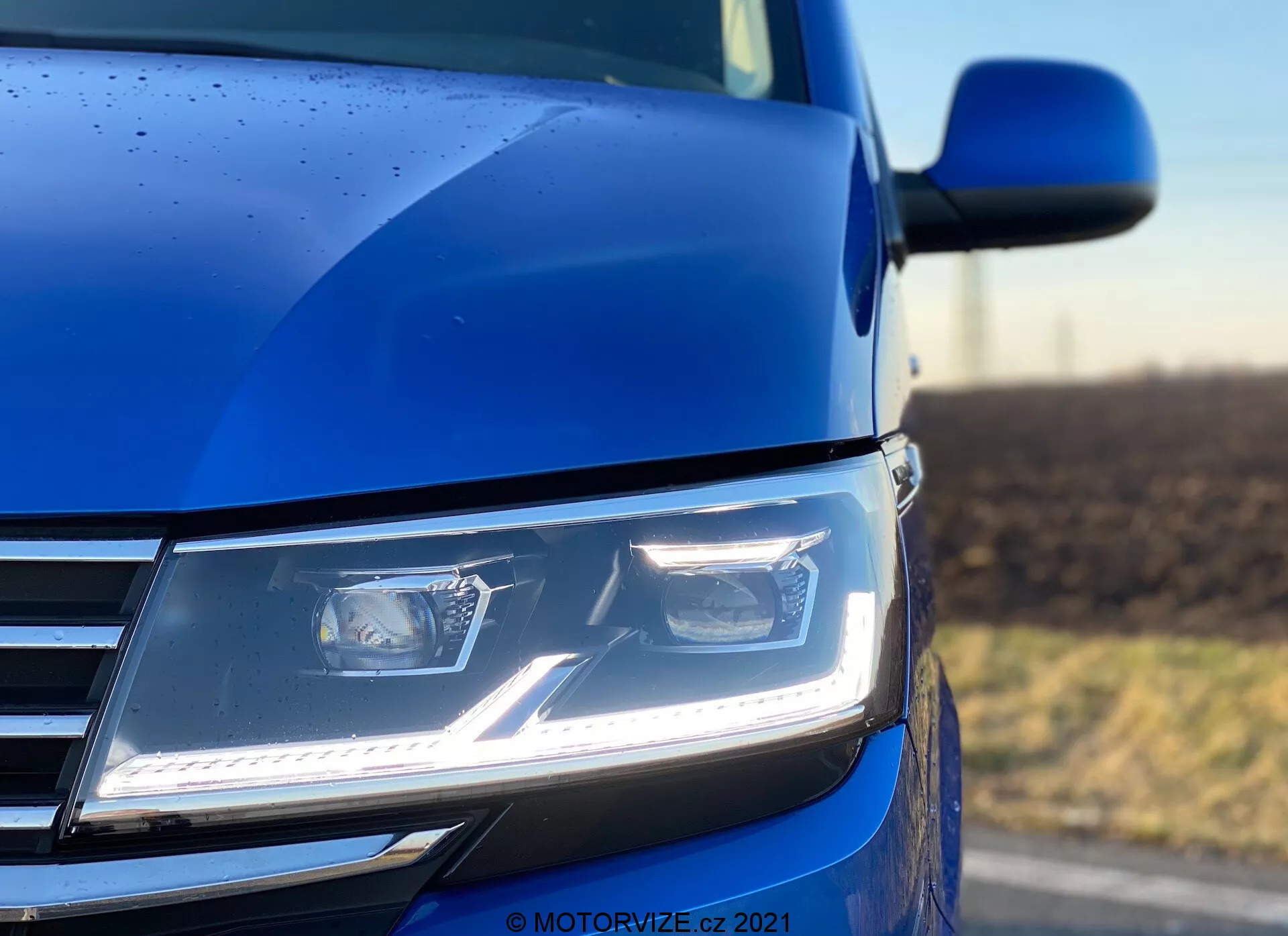 A close-up, low-angle shot of the front left section of the 2019 VW Nutzfahrzeuge Transporter (T6.1 Multivan) in blue, featuring the sharp and angular modern LED headlights with daytime running lights, part of the chrome-accented grille, and a glimpse of the hood contour and side mirror. The photo, taken with a shallow depth of field, emphasizes the headlight and surrounding bodywork, highlighting the vehicle's advanced lighting technology, contemporary design, and paint finish quality, complemented by rain droplets indicating a detailed exterior coating.