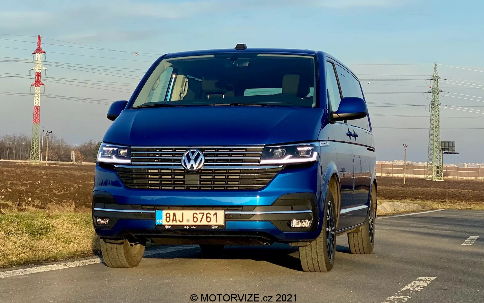 Blue Volkswagen T6-1 Multivan 2019 captured from a low front angle highlighting grille, LED headlamps with daytime running lights, Volkswagen emblem, wide grille, two-tone bumper, and alloy wheels; parked by a road with clear sky, distant foliage, power pylon, and infrastructure in the background.