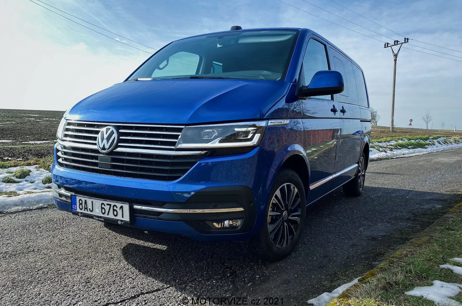 Blue Volkswagen T6.1 Multivan Transporter 2019 captured from a low front three-quarter angle, showcasing the car's front fascia, grille with prominent Volkswagen logo, horizontal chrome bars, angular LED headlights with daytime running lights, front bumper with integrated fog lights and air intakes, dynamic creased line extending along the sides, multi-spoke alloy wheels, integrated side mirror turn signals, tinted windows. The vehicle is parked on a wet road beside snowy grass, with a rural background featuring utility poles and open skies.
