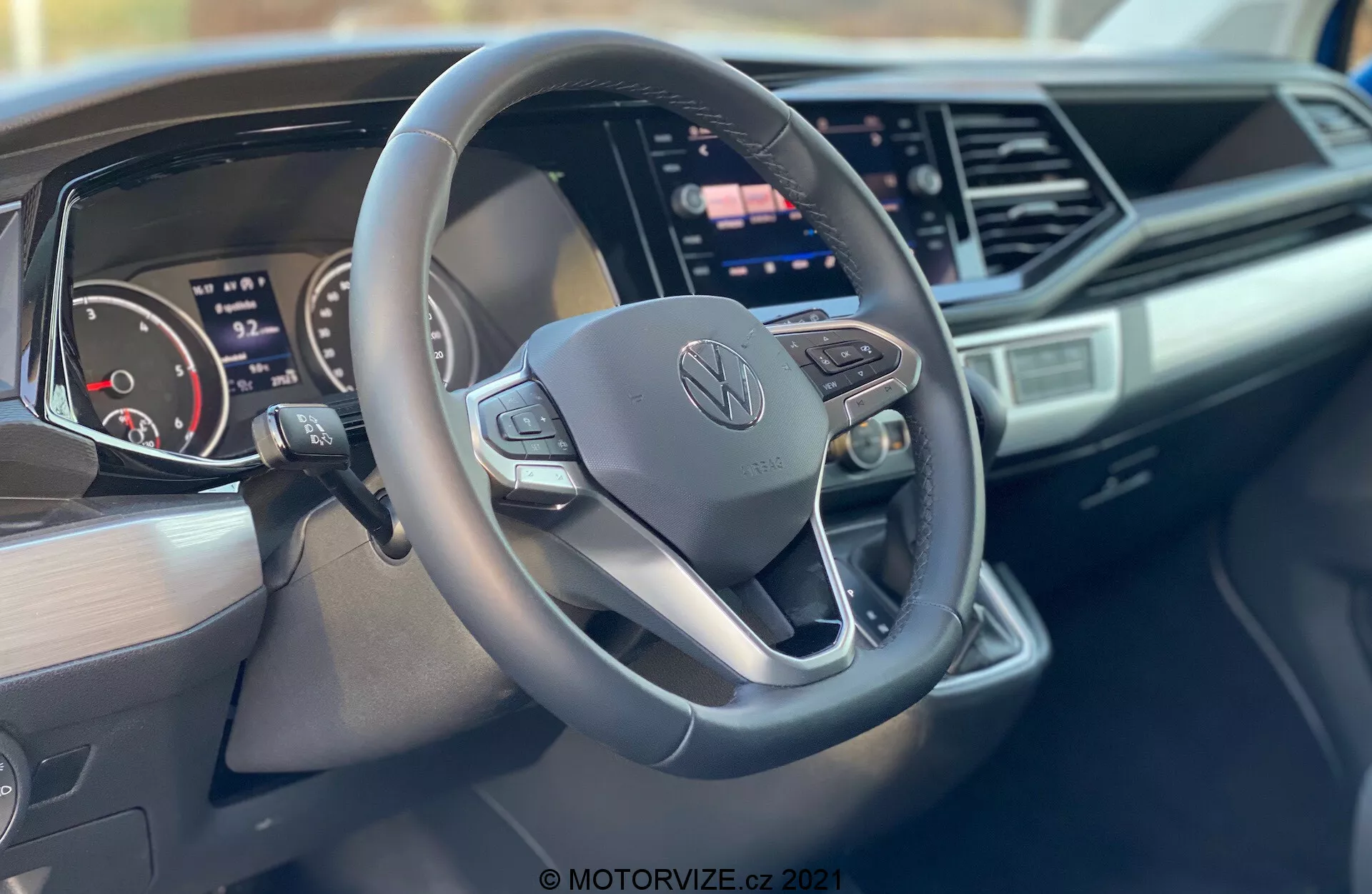 Interior shot of a 2019 Volkswagen Nutzfahrzeuge Transporter (T6-1 Multivan) focusing on the steering wheel and dashboard. The close-up, taken from the passenger side, highlights a three-spoke, flat-bottom steering wheel with control buttons and a central Volkswagen logo, a digital instrument cluster, a large central infotainment touchscreen, sleek dashboard design with angular accents, and branding on the passenger side of the dashboard.