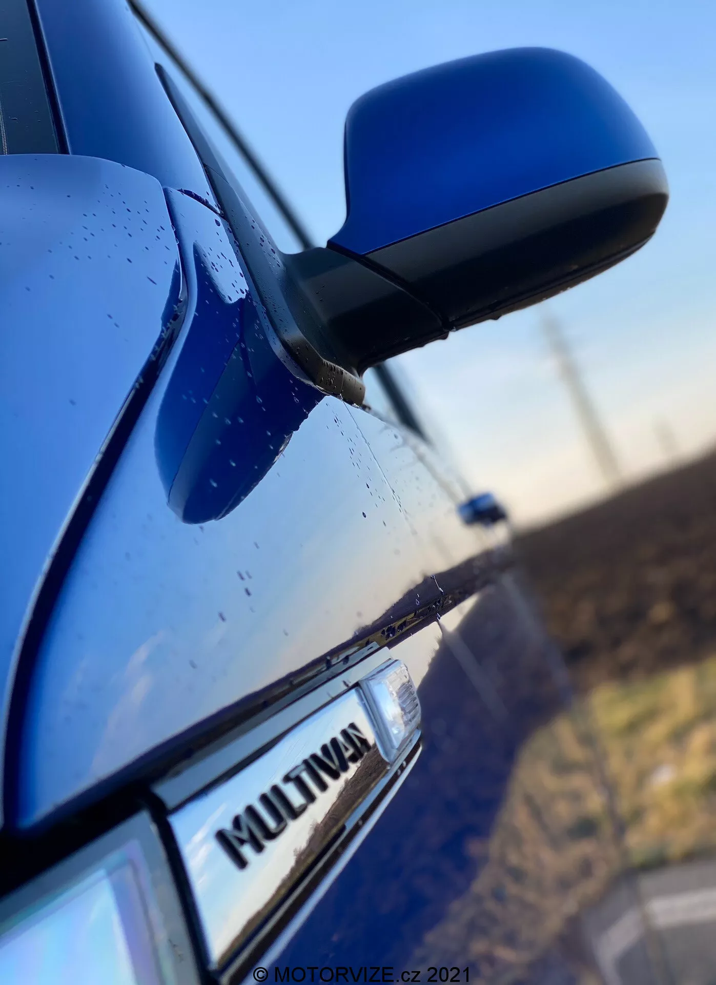 Close-up shot of the side of a blue VW Nutzfahrzeuge Transporter 2019 (T6-1-Multivan) featuring part of the front passenger door, side mirror, and a portion of the rear door. The focus is on the 'MULTIVAN' badge on the car, with the sleek and aerodynamic side mirror slightly blurred. The mirror housing matches the car body color, and water droplets are visible on the surface, suggesting weatherproof characteristics or recent rain.