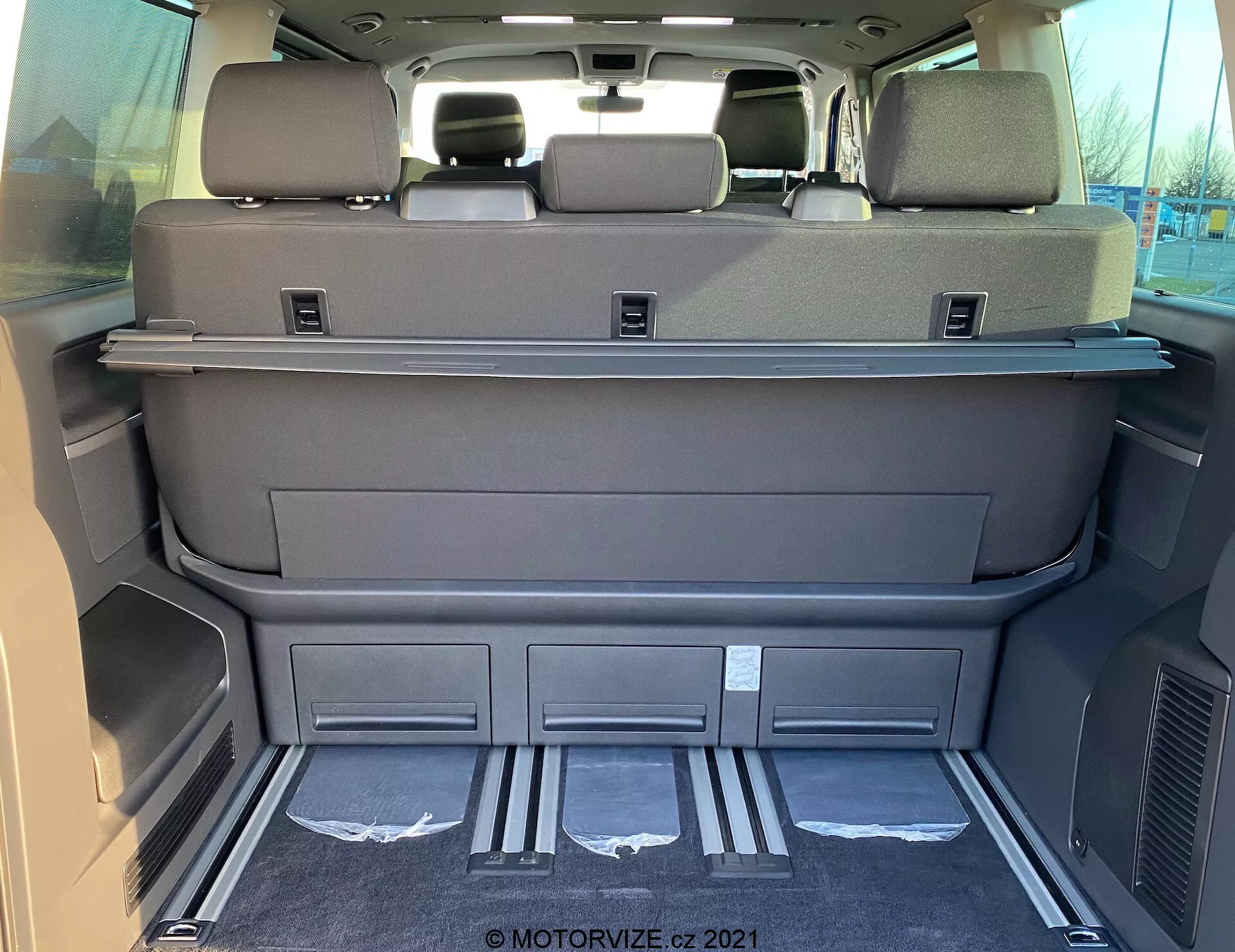 Interior view of a 2019 VW Nutzfahrzeuge Transporter T6.1 Multivan cargo space from the rear, showing spacious area with folded third-row seats, dark grey or black color, cargo cover shelf, headrests on rear seats, metallic entry guards, molded compartments, air vents, clean carpeting, and possible electronic seat or tailgate controls.