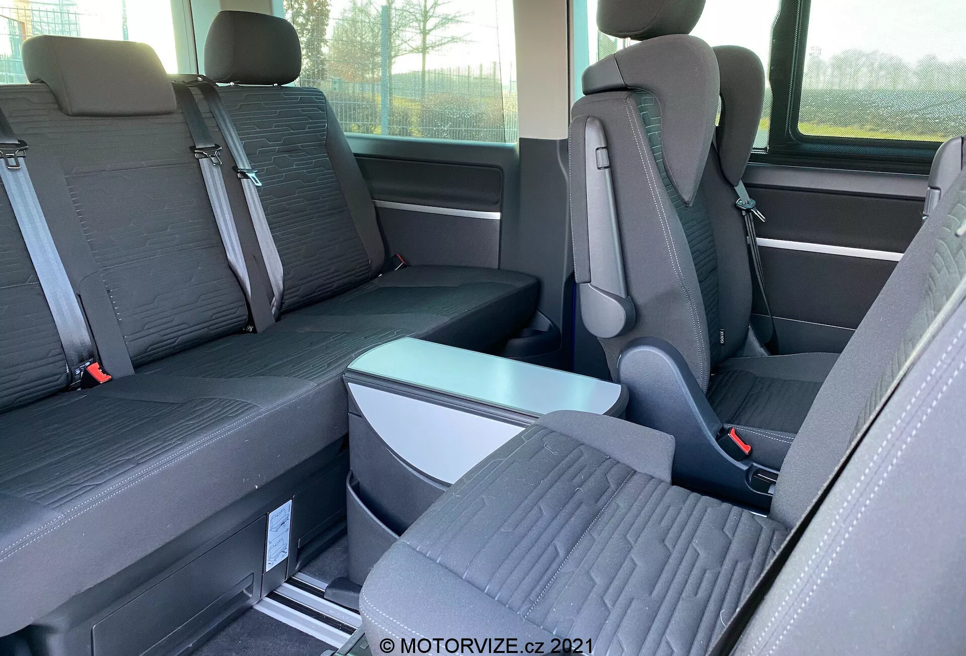 Interior view of VW-Nutzfahrzeuge Transporter 2019 (T6.1 Multivan) focusing on the second and third-row seats. Photographed from the side at eye level, the image shows individual seats with prominent headrests in the second row and a three-passenger bench seat in the third row. The seats are two-tone fabric, and the floor features metal tracking for reconfigurable seating. A foldable armrest and seat belt buckles are visible. Partial window and side storage are also seen.