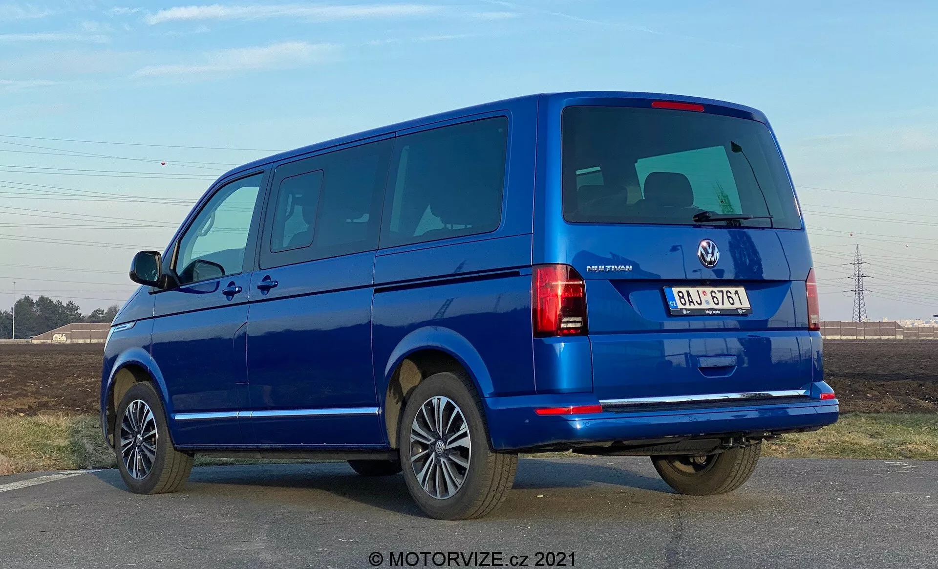A blue VW-Nutzfahrzeuge Transporter 2019 (T6-1 Multivan) viewed from a three-quarter rear angle. The image captures the rear light clusters, rear windows, and tailgate as well as the side profile with passenger doors and windows. The van occupies a large portion of the frame at eye level, revealing split rear doors and several side windows, indicating a passenger model. Alloy wheels, body-colored bumpers, and side mirrors suggest a high trim level. The lighting suggests daytime with clear skies, and there are crisp shadows under the vehicle.
