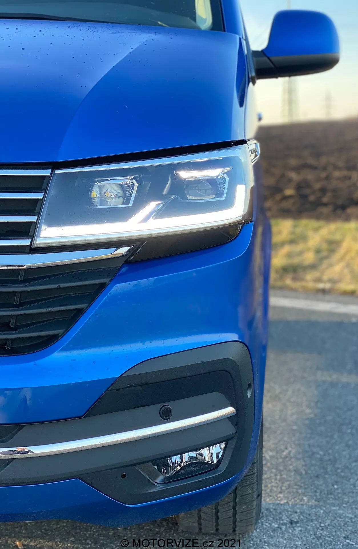 Close-up of the front-left side of a 2019 VW Nutzfahrzeuge Transporter (T6.1 Multivan) highlighting the metallic blue paint, advanced LED headlights, modern grille design with horizontal lines, and a sophisticated front bumper featuring fog lights or sensor units. The photo angle emphasizes the car's aerodynamics and design aesthetics.