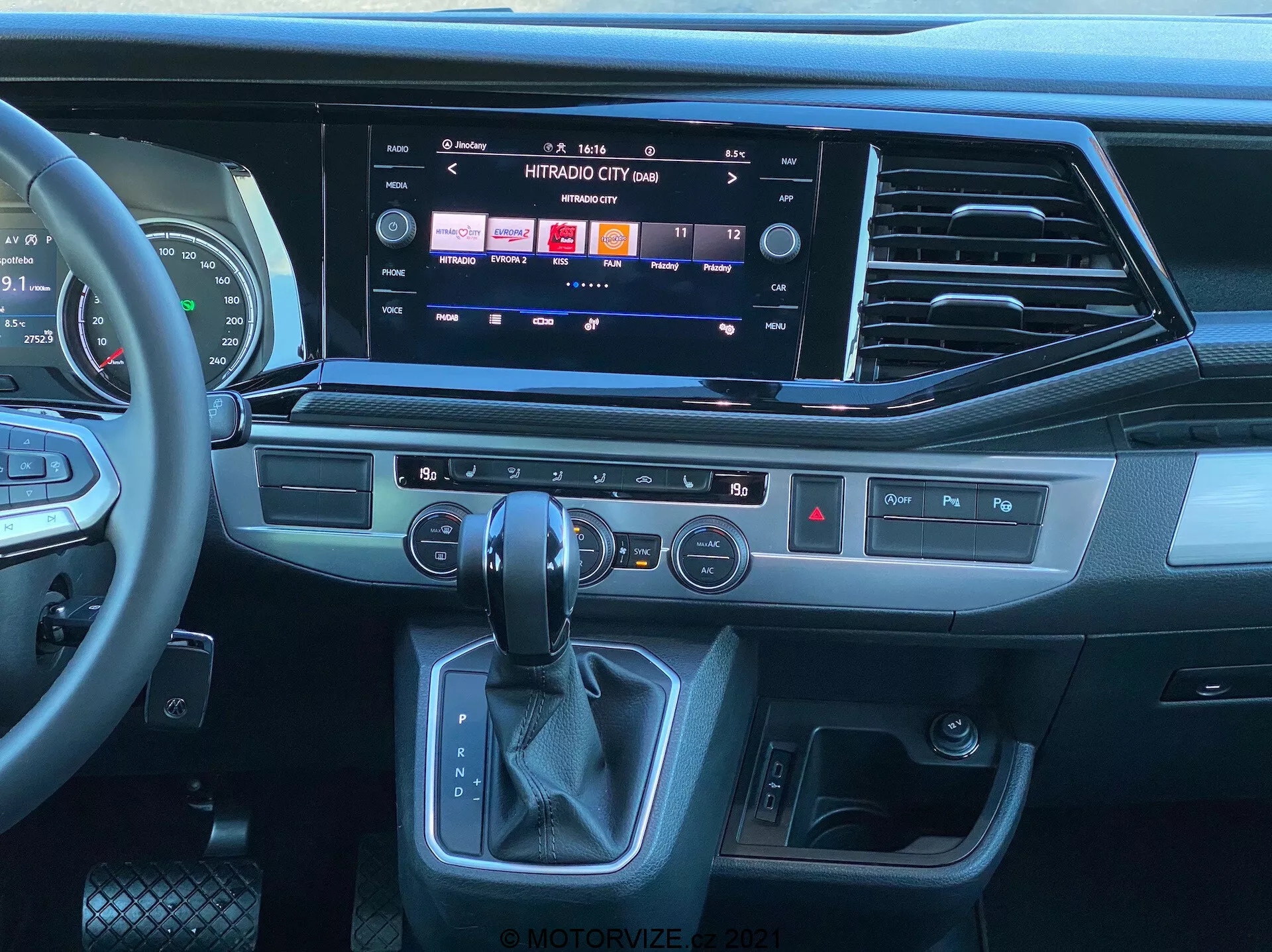 Interior view of a 2019 VW Nutzfahrzeuge Transporter T6.1 Multivan from the passenger seat perspective, showcasing the modern dashboard and center console. Multifunctional leather-wrapped steering wheel with media controls, large touchscreen infotainment system displaying media and connectivity options, sleek air vents, tactile buttons for quick functions, dual-zone climate control panel, ergonomic automatic gear shift lever area, and keyless start/stop ignition button. The design features a mix of dark plastics, brushed metal-like trim, and leather-like materials, highlighting a contemporary, premium feel.