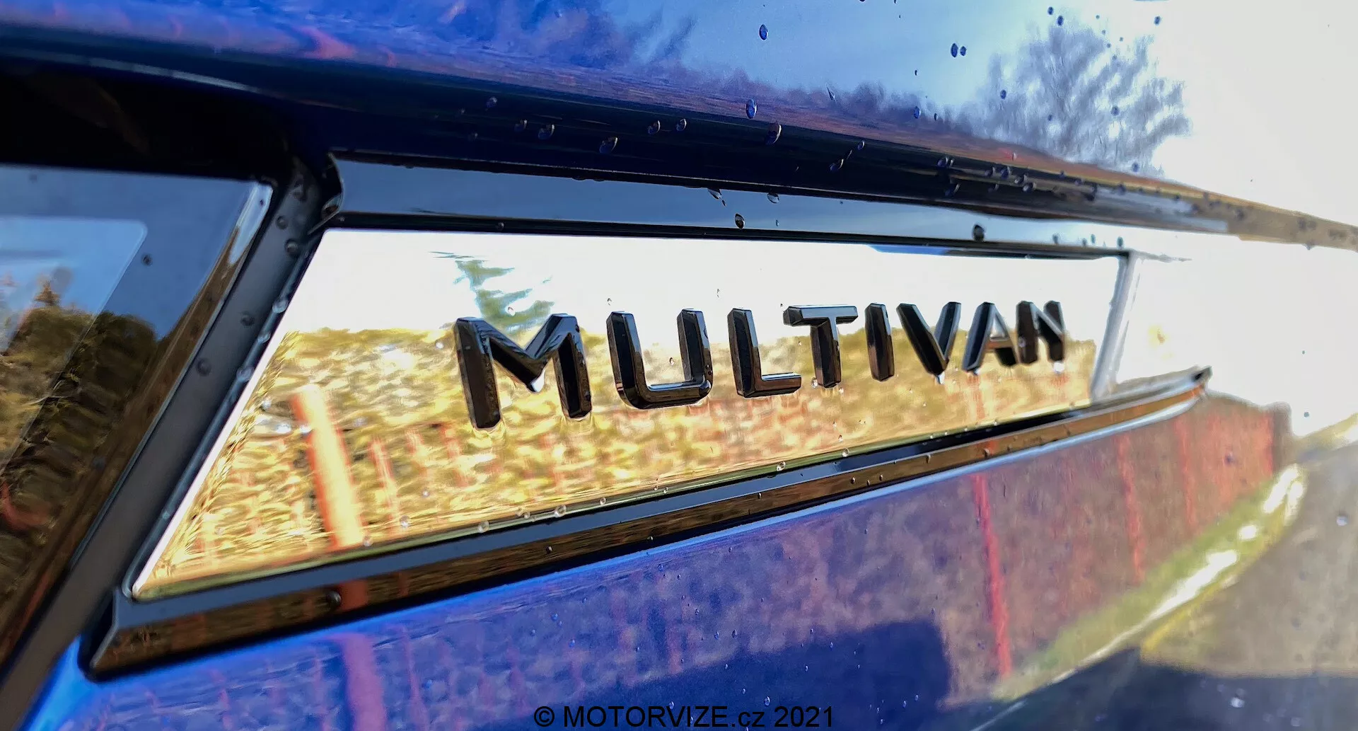 Close-up view of part of the VW Nutzfahrzeuge Transporter 2019 (T6-1 Multivan) focusing on the 'MULTIVAN' badge. The image is taken from a low rear side angle, looking slightly upwards where the rear quarter window meets the edge of the roof. Slight reflections on the window's glass suggest an outdoor setting. This tightly cropped image emphasizes the manufacturer's model designation emblem.
