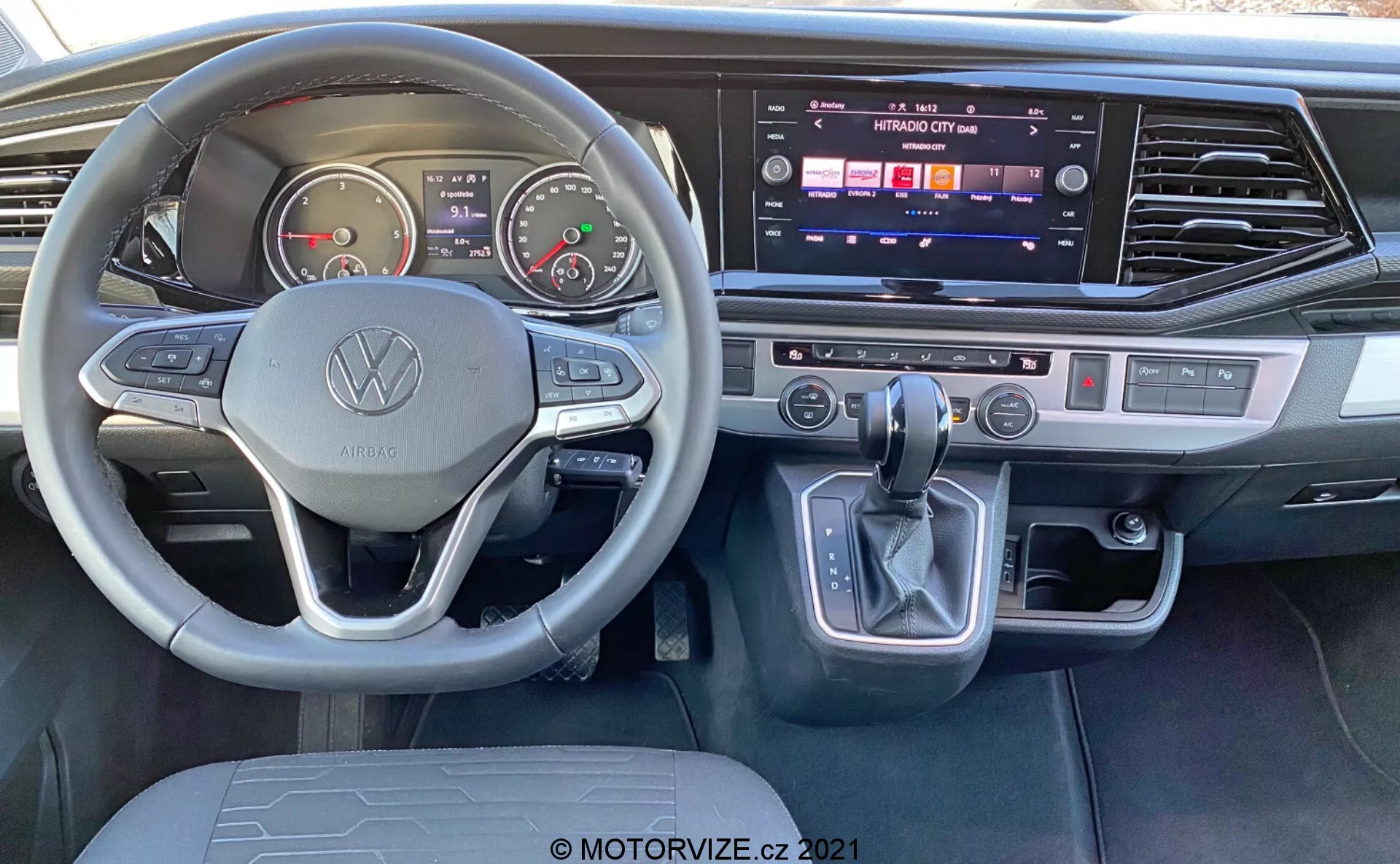 Interior view of the 2019 VW Nutzfahrzeuge Transporter T6.1 Multivan from the driver's seat perspective. Features a Volkswagen-branded steering wheel with multifunctional buttons, analog gauges, digital display, center console with active touchscreen infotainment system, dual-zone climate control, gear shift lever with electronic park button, and modern brushed metal accents.