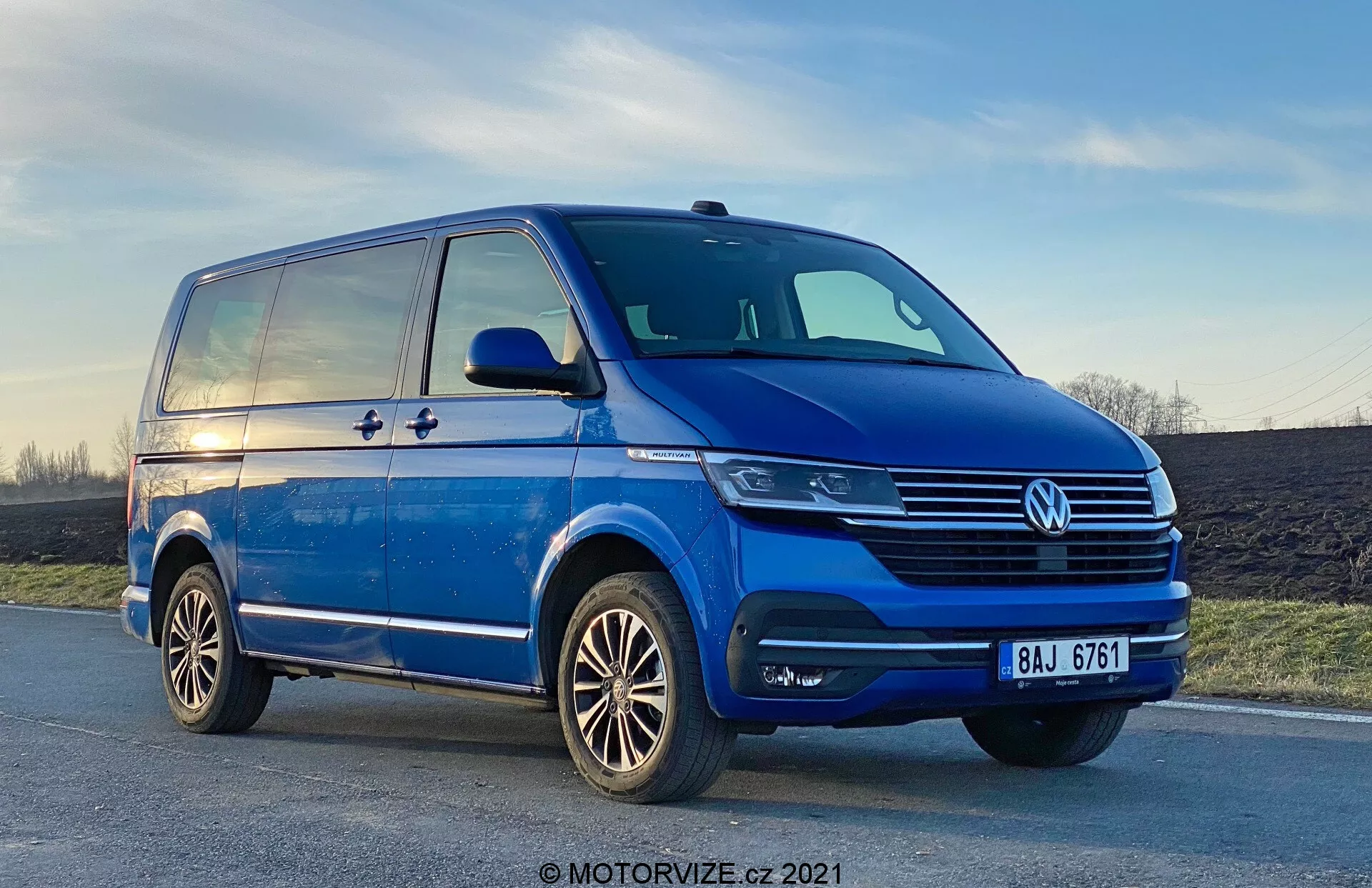 Low-angle view of a blue Volkswagen T6-1 Multivan 2019, showcasing the front left quadrant from a three-quarter perspective. The van is parked on a slightly inclined road, with prominent chrome detailing on the grille, a central VW emblem, and modern headlamp units potentially featuring LED technology. Fog lights are integrated into the bumper, which also has air intake vents. The side profile reveals a sliding door, a character line extending from front to rear, and substantial side mirrors with turn indicators. Alloy wheels with a multi-spoke design and well-maintained tire treads are also visible. The sky in the background shows a hint of sunset.
