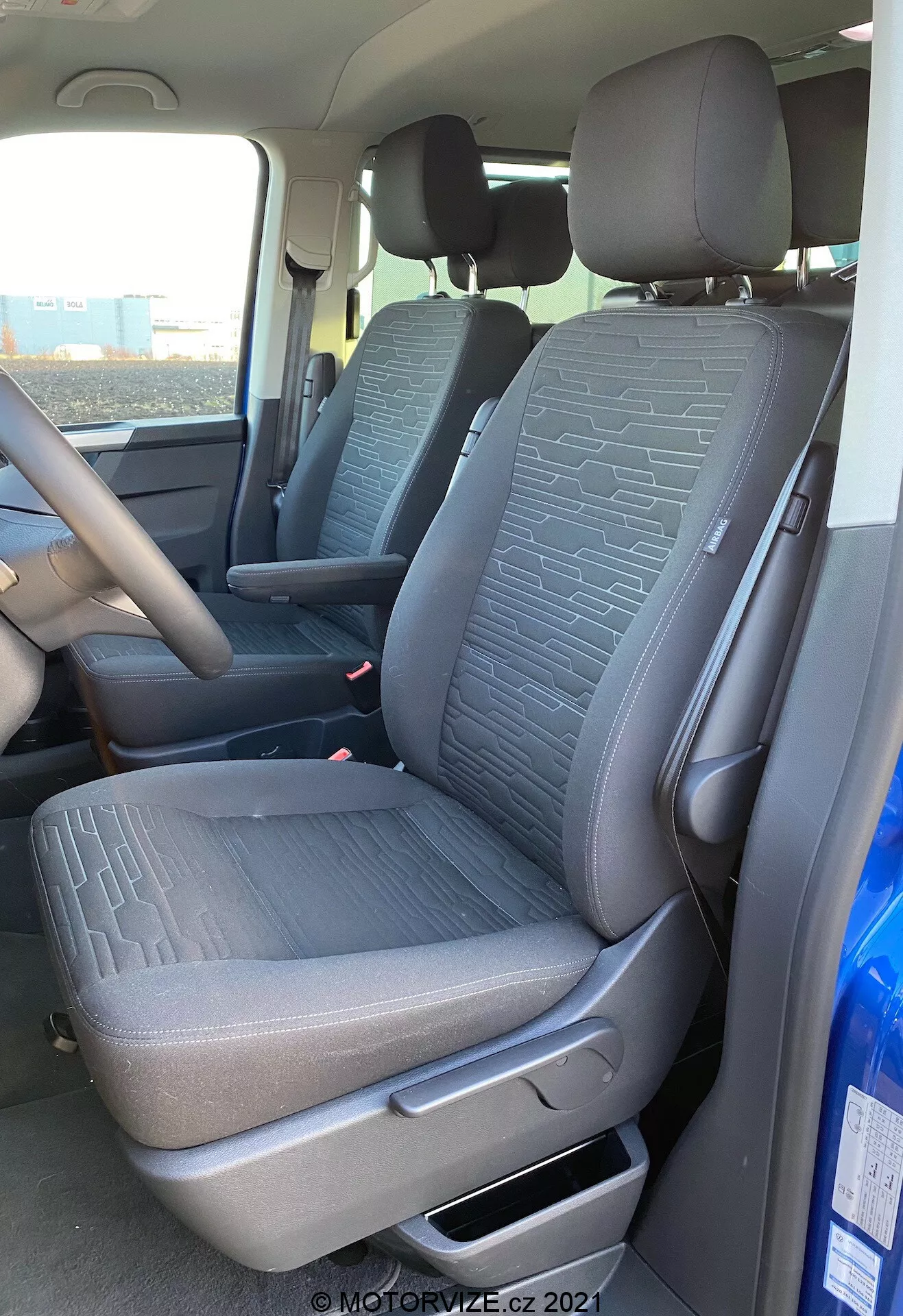 Interior of a 2019 VW Nutzfahrzeuge Transporter T6.1 Multivan, showing front passenger seat and part of driver’s seat from an exterior passenger side perspective. The shot, taken at a slightly low angle, highlights the textured fabric with geometric patterns on the seats, manual adjustment lever, seatbelts, and part of the center console and steering wheel. Natural daylight illuminates the vehicle's interior.