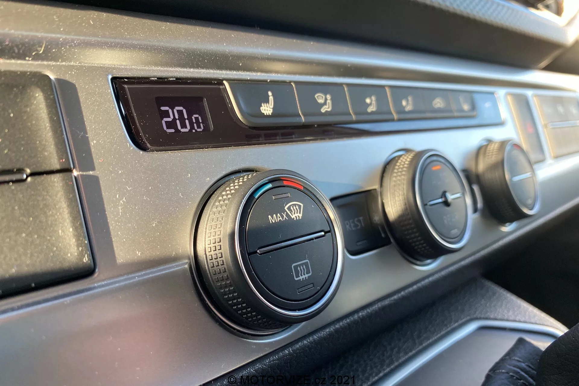 Close-up view of the climate control panel on the dashboard of a 2019 VW Transporter T6.1 Multivan from the perspective of a front-seat occupant, featuring a digital temperature display set to 20.0 degrees Celsius, airflow direction controls, three rotary dials for air conditioning and fan settings, and a recirculation button, all with a textured, black plastic rim and a smooth, dark dashboard finish.