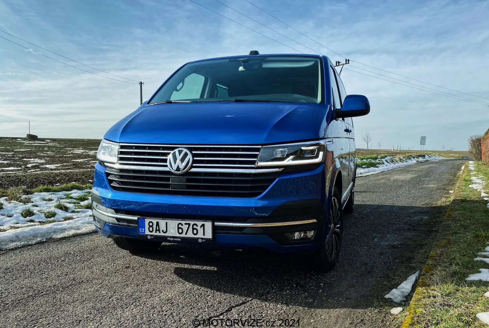 A low front three-quarter view of the Volkswagen T6.1 Multivan 2019, featuring the front grille with horizontal lines, distinctive VW emblem centered on the grille, headlights possibly with LED DRLs and bi-xenon or LED lights, and front bumper with fog lights and air intakes. The image showcases part of the left side including the driver’s door and fender, with the hood and windshield visible. The car is deep blue and parked on a rural road with snow on the ground and a clear blue sky above.