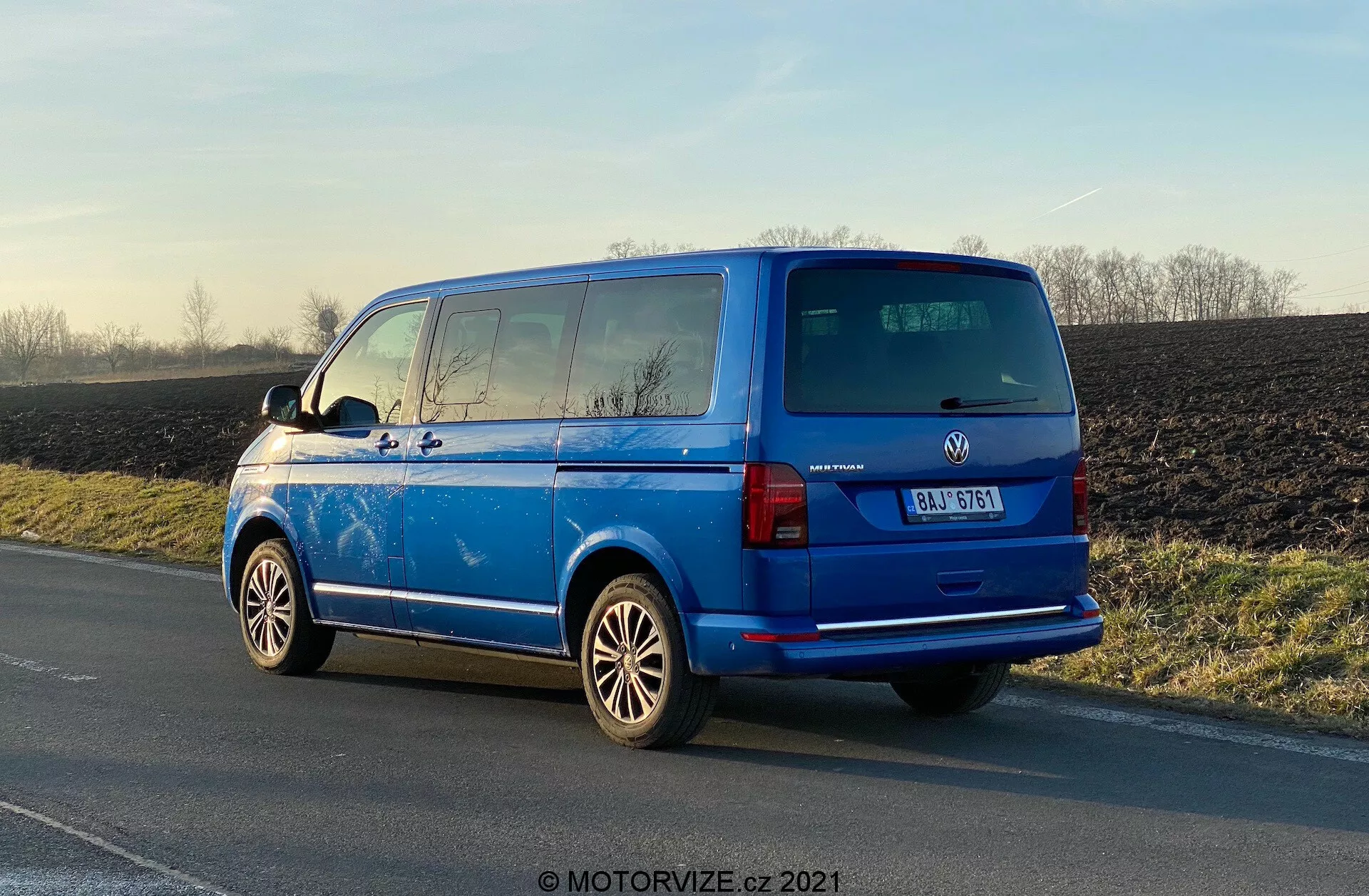 Vibrant blue Volkswagen Transporter 2019 (T6-1 Multivan) parked on road shoulder, rear three-quarter view showing tail lights, rear window, back door, manufacturer badge, vehicle model details; prominent alloy wheels, external side mirrors, rear window defroster lines, privacy-tinted second-row windows, roof antenna; parked in natural daylight with glare highlighting polished surface, rural background.