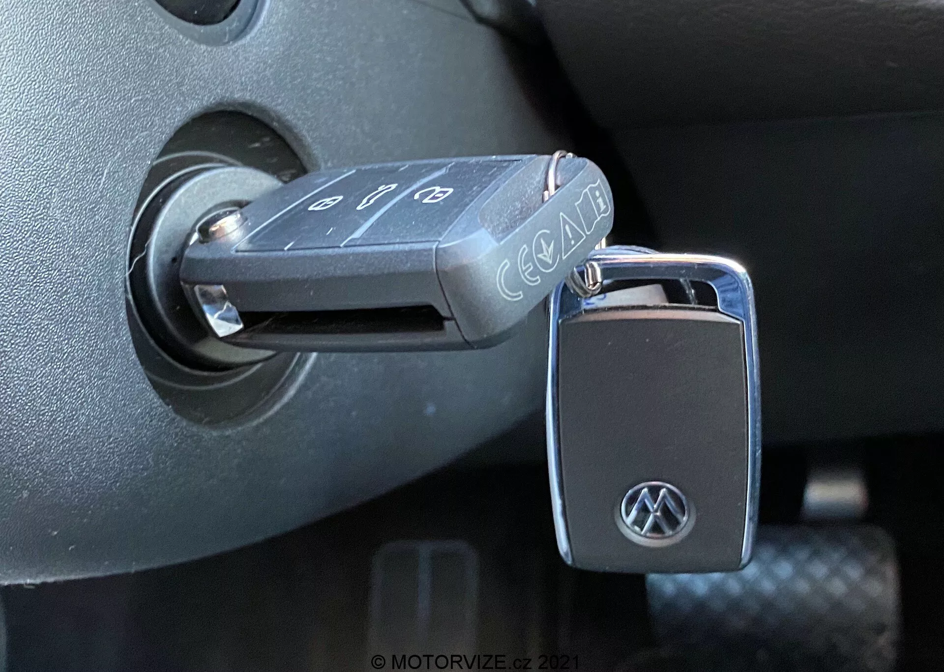 Close-up of a Volkswagen flip key inserted into the ignition switch of a 2019 VW Nutzfahrzeuge T6-1 Multivan. The key, with buttons for door lock, unlock, and panic alarm, shows a VW logo on the fob. The ignition switch is located on the steering column's dark plastic surroundings, indicating the ignition is in the 'off' position. The photo, taken from a side-on angle with natural light, highlights the detailed textures and finishes of the vehicle's interior.