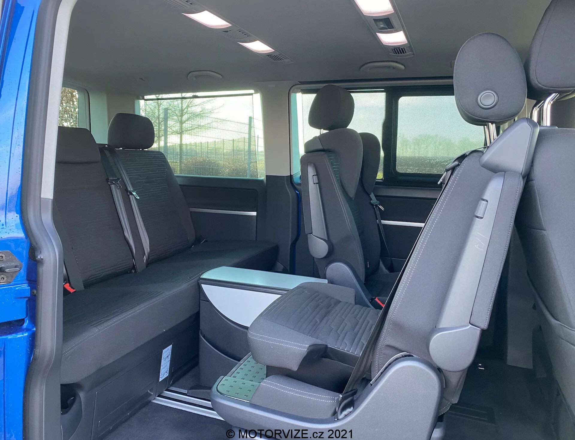 Interior of 2019 VW Transporter T6.1 Multivan showing the second and third row seats from the rear passenger-side perspective, featuring a 2-3-2 seating configuration with dark fabric upholstery. Partial exterior view through open door, headrests on all seats, and elevated third-row seats for improved visibility.