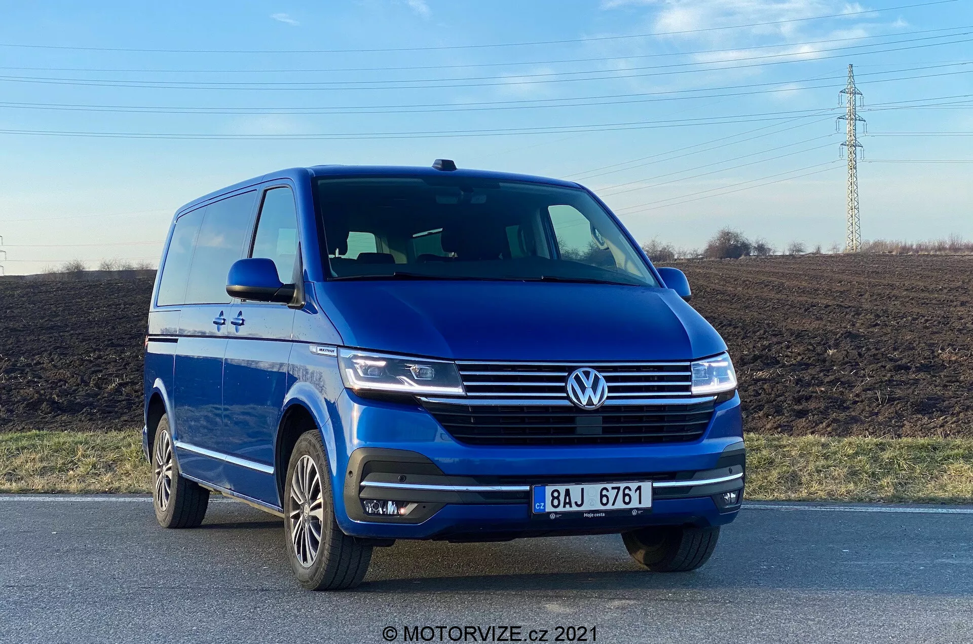 The image features a blue 2019 Volkswagen VW-Nutzfahrzeuge Transporter (T6-1 Multivan) captured from a low frontal angle, showcasing the front left quarter with a clear view of its front fascia and driver-side profile. The vehicle is on a road, highlighting elements like the prominent Volkswagen badge, sleek headlights, sculpted hood lines, integrated fog lights on the bumper, and multi-spoke alloy wheels. The photograph, taken in daylight, emphasizes the van's exterior styling and driver-assistance technologies such as parking sensors.