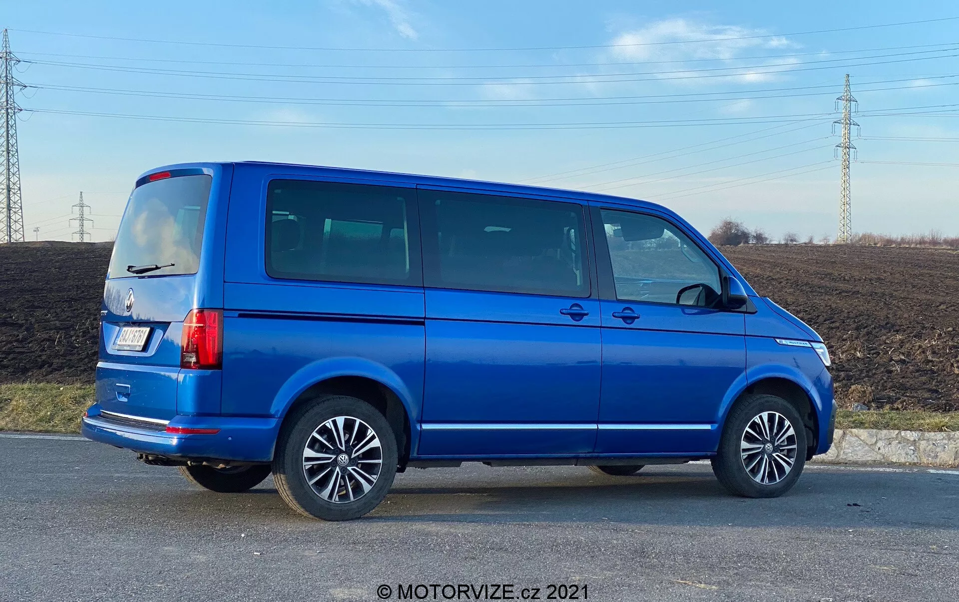 A full side profile view of a blue 2019 VW Nutzfahrzeuge Transporter (T6.1 Multivan) with alloy wheels, visible door lines, tinted side windows, boxy silhouette, vertical tail lights, roof antenna, and bright blue metallic paint. The image is taken at an angle, providing perspective on both the side and rear portions of the vehicle, set in a rural area with a field and power lines in the background, under daytime lighting.
