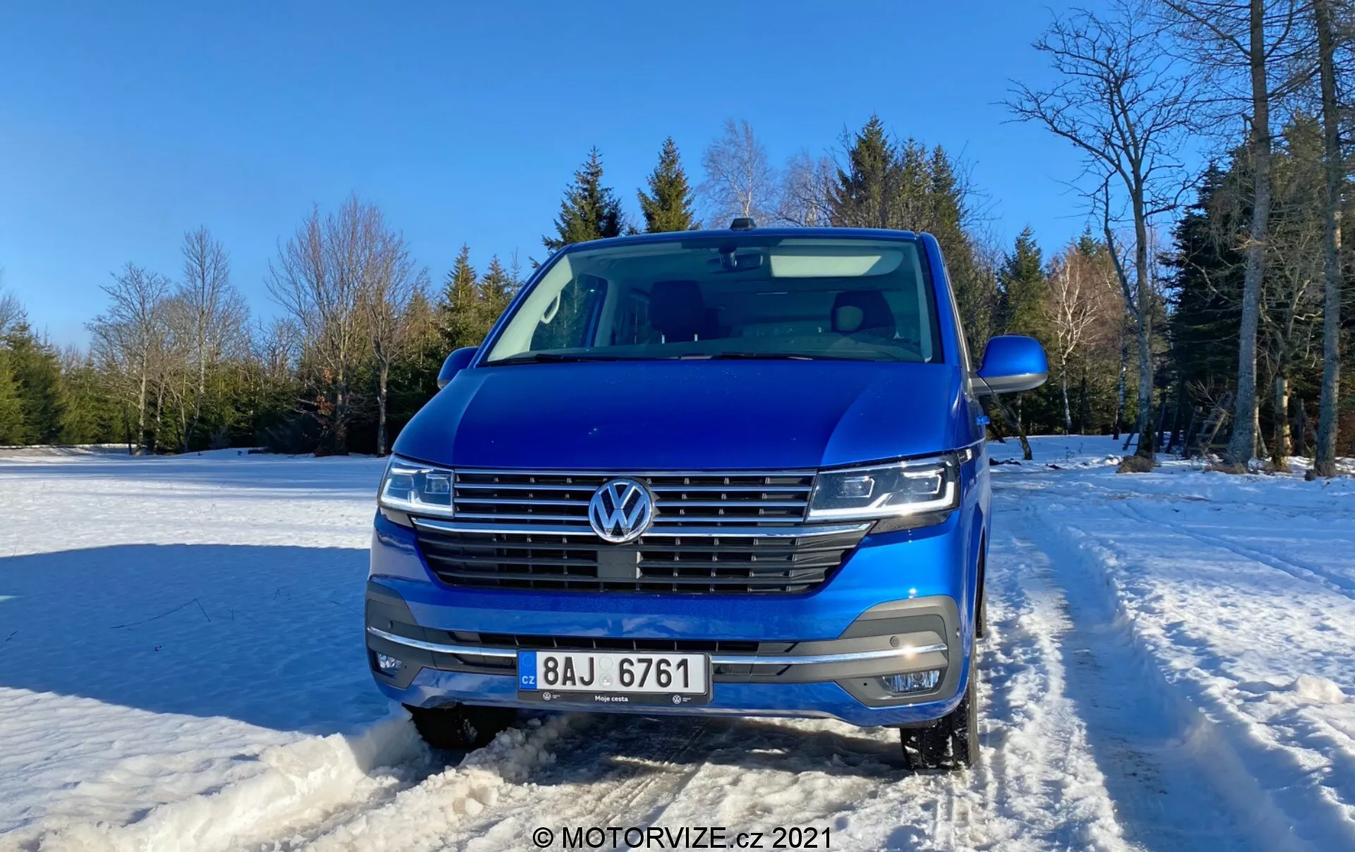 Frontal shot of a blue 2019 Volkswagen Transporter (T6-1 Multivan), showcasing its grille, headlights, and hood area. Positioned outdoors on snow-covered ground with clear sky and trees in the background. Car features include VW emblem, chrome-trimmed horizontal grille, LED headlights, front-facing camera, parking sensors, silver roof rails, and partially visible side mirror. Contemporary design with streamlined front fascia, photographed in daytime with soft sunlight from right side.