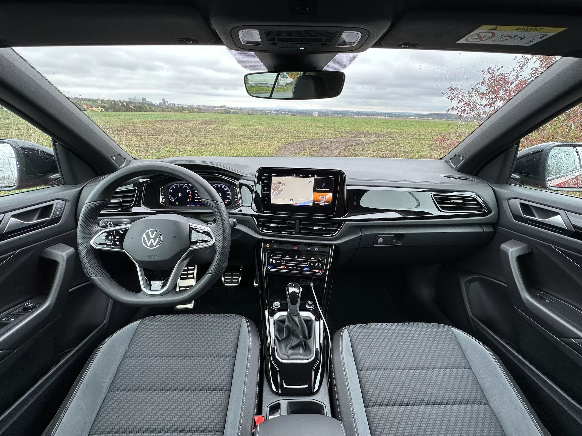Interior view of the 2022 VW T-Roc Cabriolet from rear to front showing streamlined dashboard with chrome accents, digital infotainment screen, advanced dual-zone climate control, leather-wrapped multi-function steering wheel with Volkswagen logo, automatic transmission gear shifter, and contemporary two-tone fabric seats with stitch pattern.