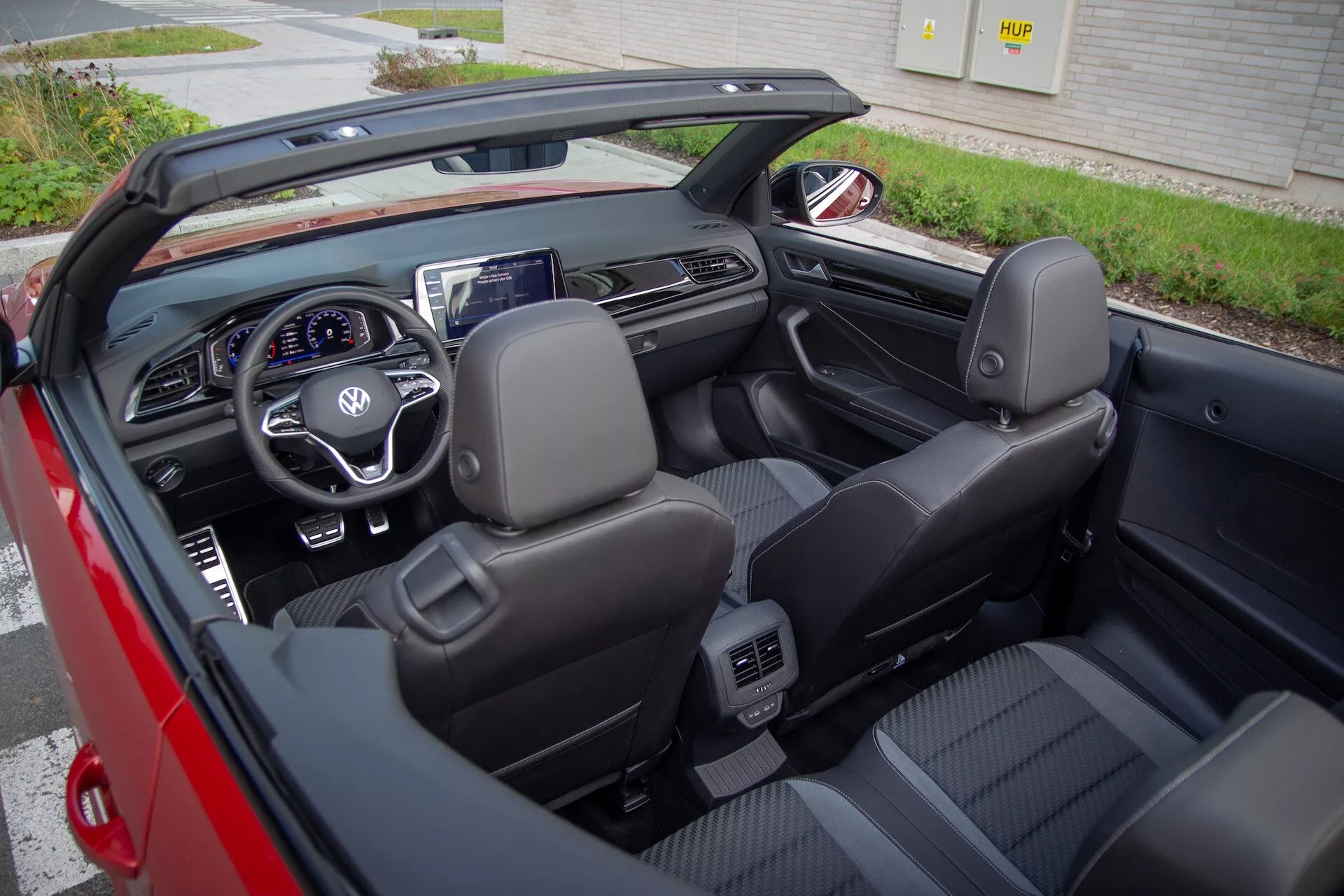 Interior view of a 2022 VW T-Roc Cabriolet convertible car with top retracted, featuring black leather seats, multifunction steering wheel, digital dashboard, and central infotainment display from an overhead slightly right-angled perspective