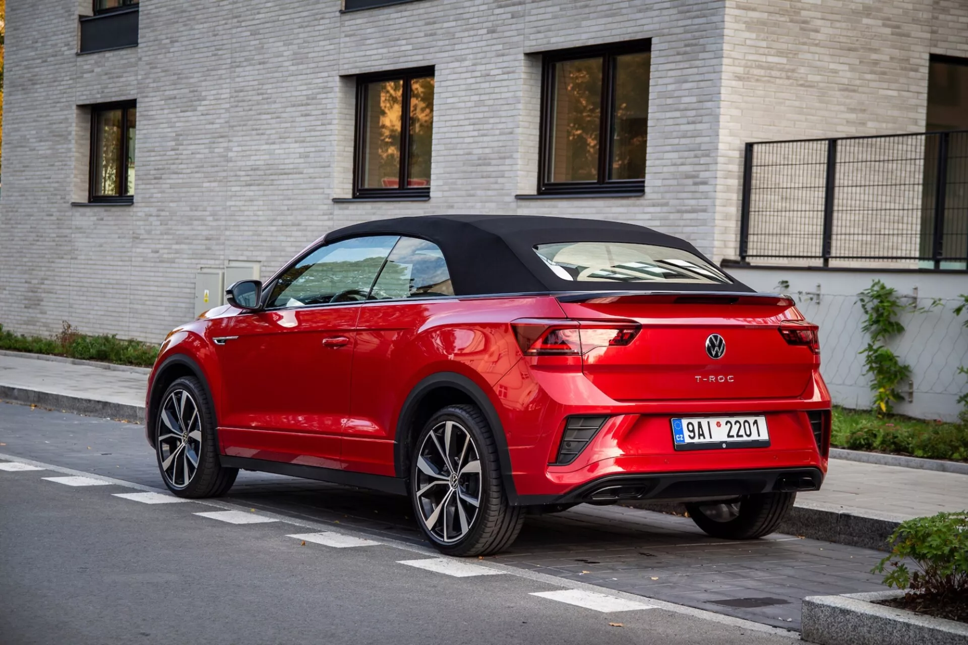 Rear three-quarter view of a red Volkswagen T-Roc Cabriolet parked on a street, highlighting its black retractable roof, sleek LED taillights, rear spoiler lip, dual exhaust tips, and integrated parking sensors against a light-colored brick building background.
