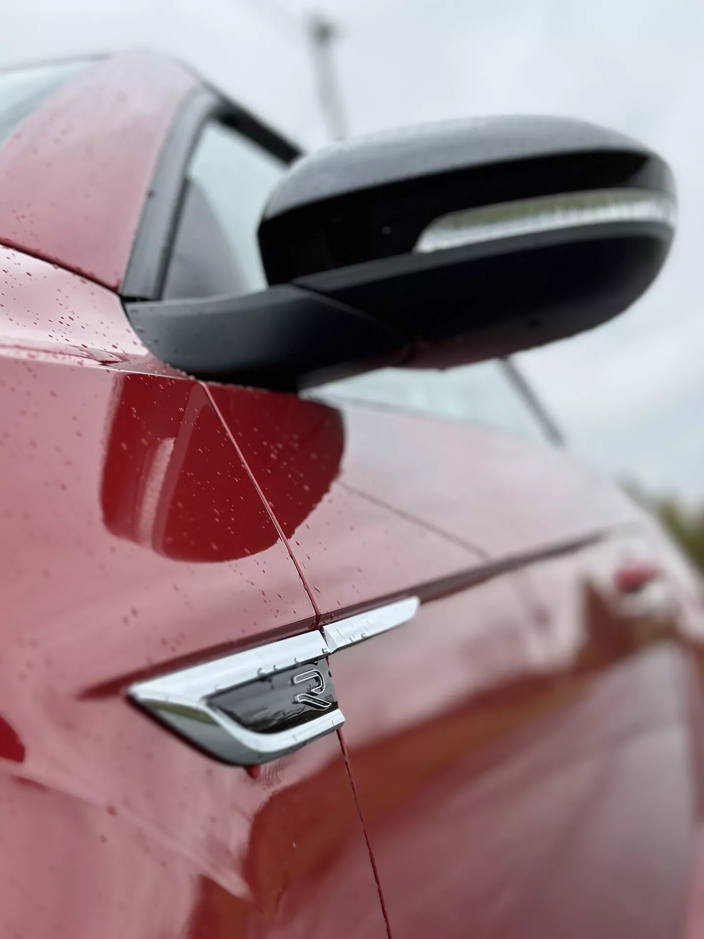 Close-up side view of a 2022 VW T-Roc Cabriolet in red, highlighting the rear part with a detailed look at the black side mirror, door handle with keyless entry, and car's design elements including badge, under a slight upward angle with water droplets on surface indicating recent rain or wash, set against a blurred background