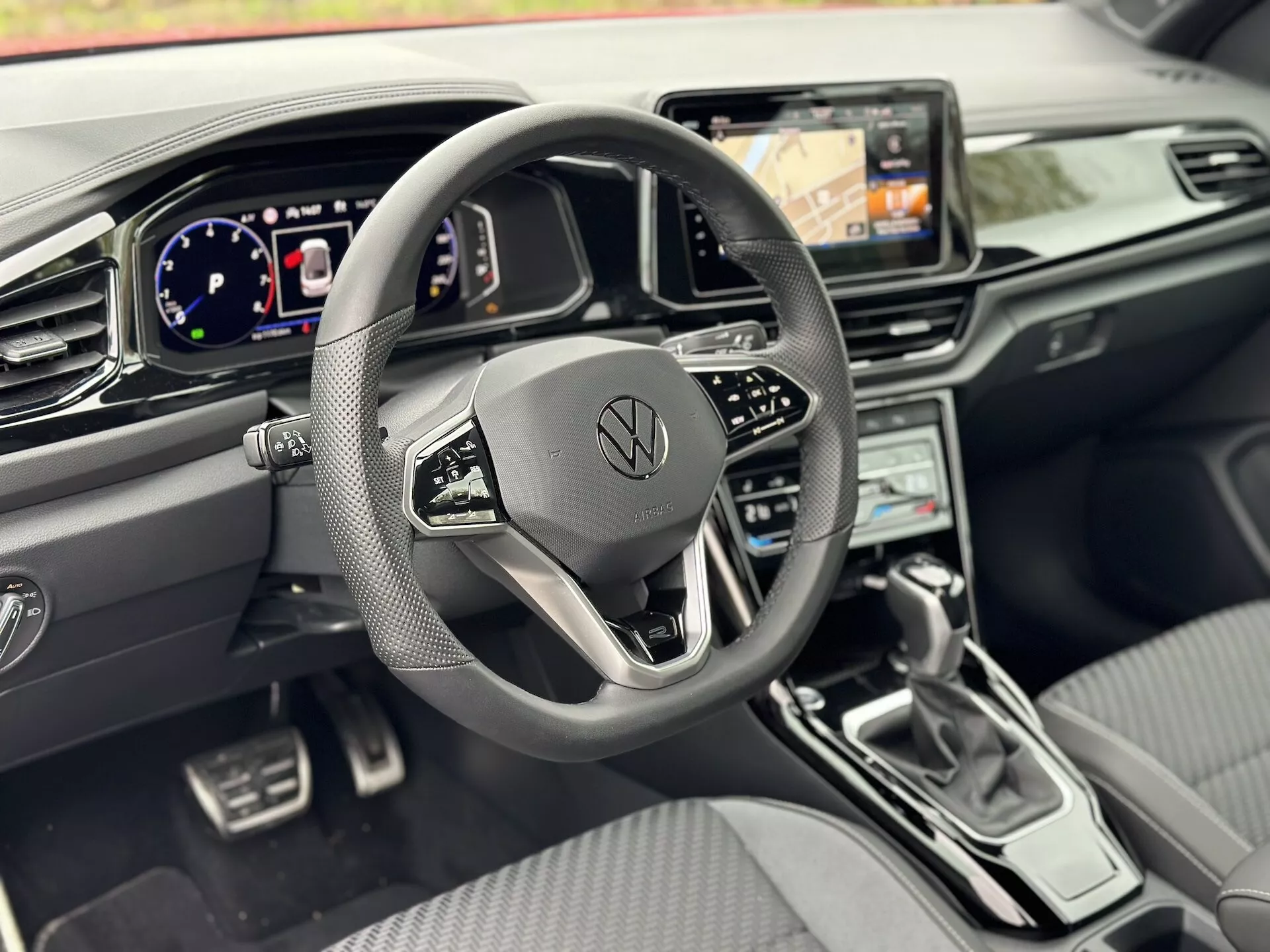 2022 Volkswagen T-Roc Cabriolet interior highlighting the driver's perspective with a focus on the three-spoke steering wheel featuring multifunction buttons and Volkswagen logo, digital instrument cluster, navigation-equipped infotainment system, climate control, sleek gear shift lever, driving assist controls, and premium metallic and leatherette cockpit design.