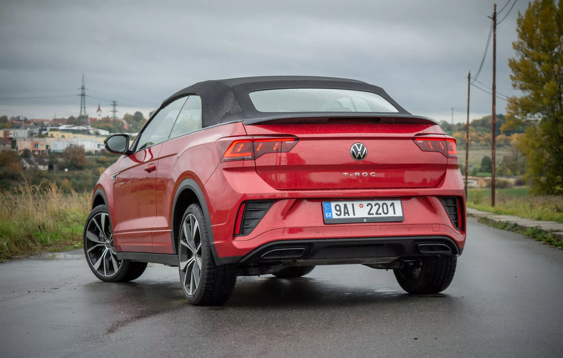 Rear three-quarter view of a 2022 VW T-Roc Cabriolet parked on a damp road showcasing its modern convertible design, sharp body lines, sporty alloy wheels, sleek rear lights, and dual exhaust, set against a semi-rural backdrop with electricity pylons and lush greenery.