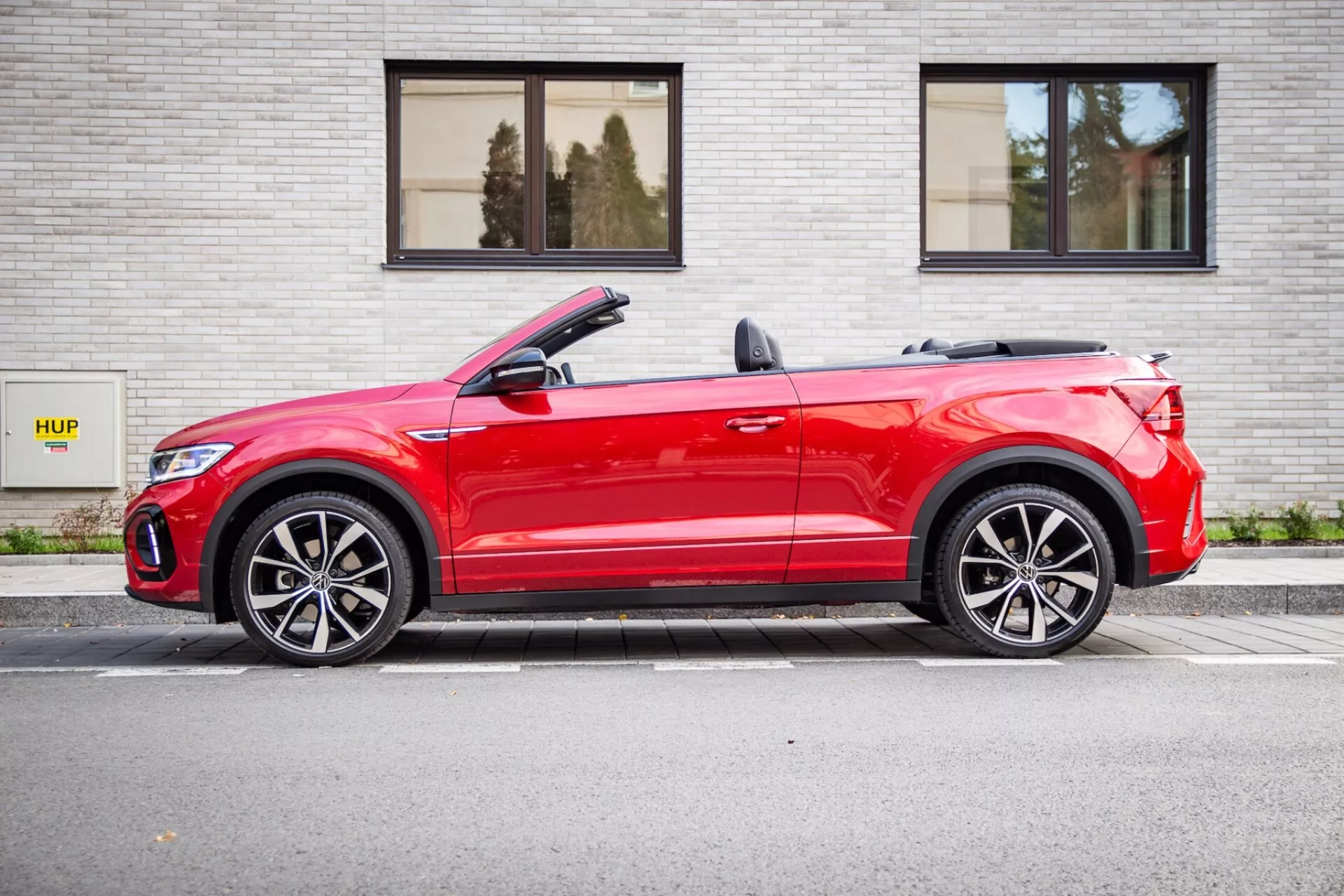 2022 Volkswagen T-Roc Cabriolet in red parked parallel to a gray brick wall with HUP sign, showing a side profile with a slight front angle, convertible top down, LED lights, and sporty alloy wheels