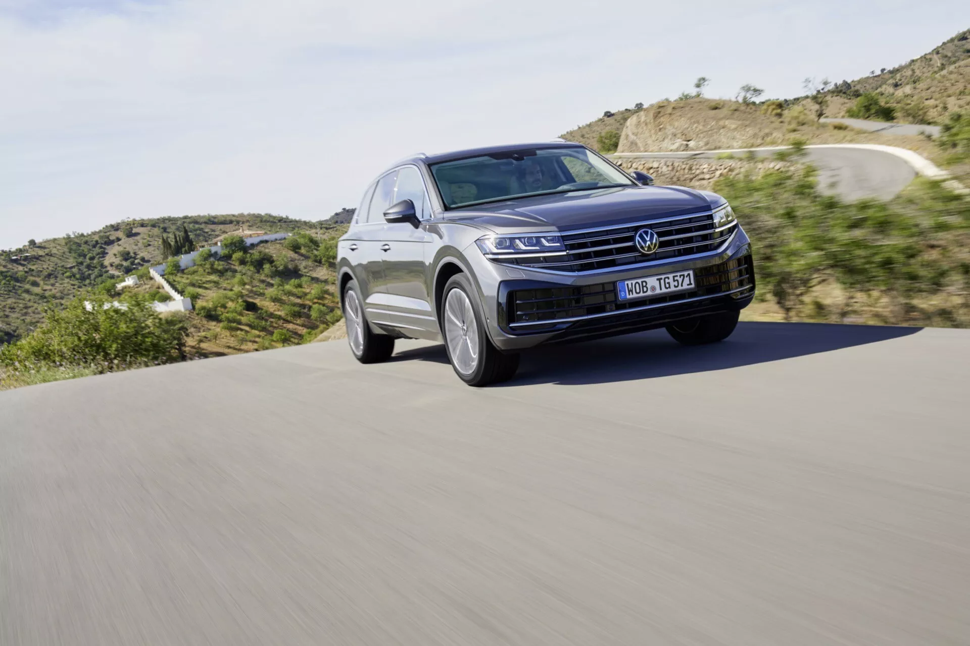 2023 Volkswagen Touareg III Facelift SUV in motion on an asphalt road, featuring a prominent front grille, LED headlamps, and the sleek contour of the hood against a natural landscape backdrop, emphasizing modern design and automotive technology.